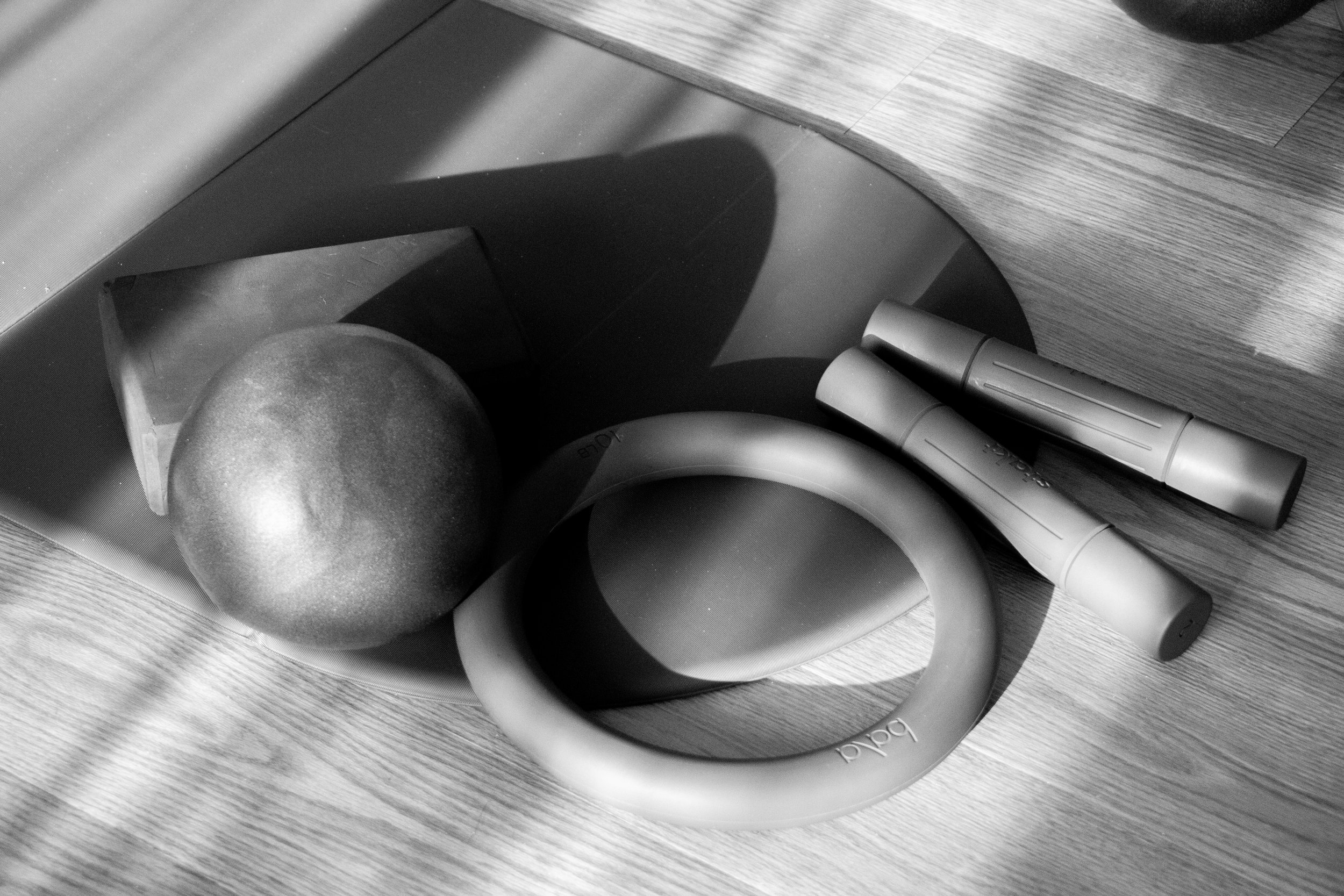 Black and white image showing a wooden floor, a portion of a chair leg, a kettlebell, a rolled-up yoga mat, and two cylindrical objects.