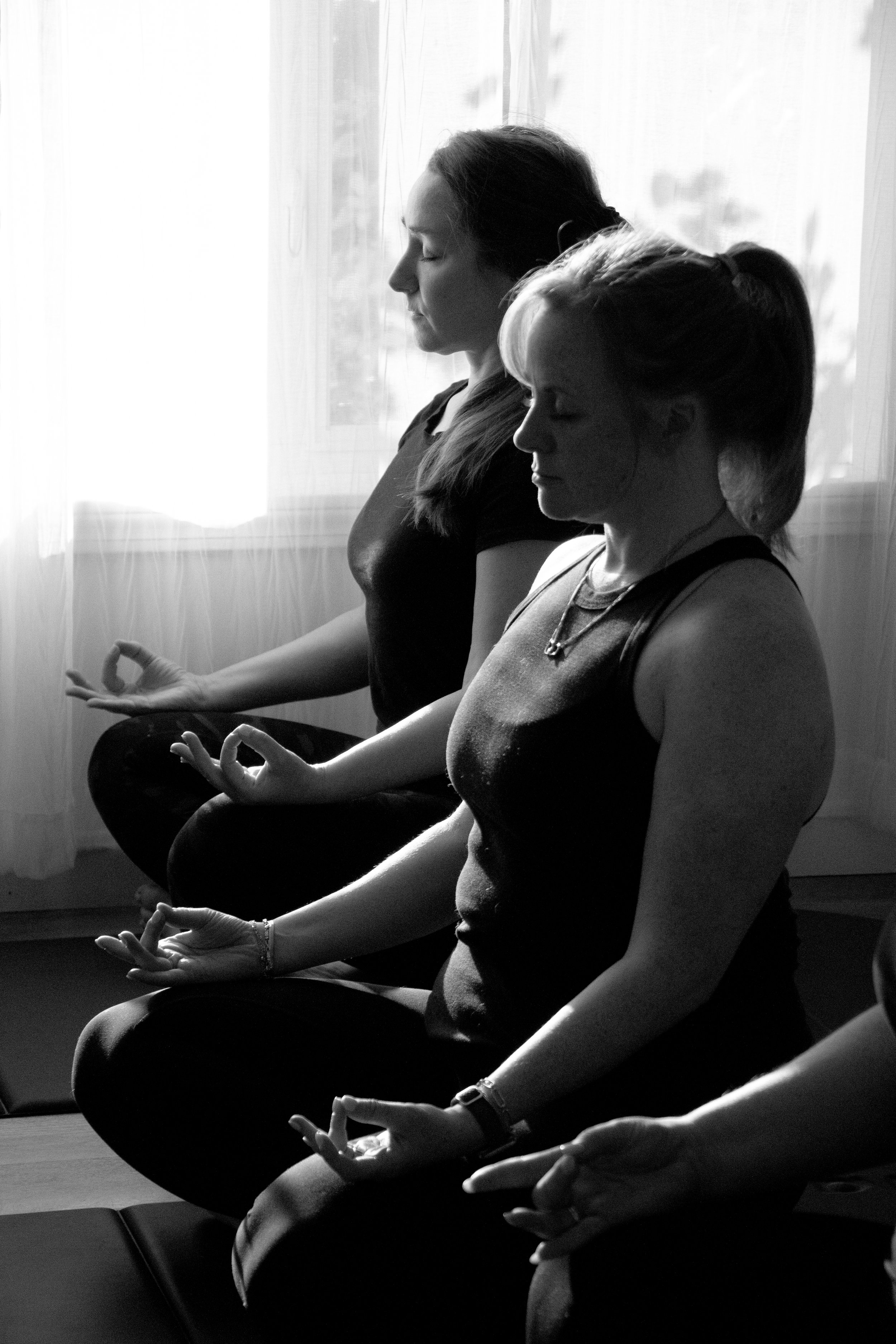 Two women practicing meditation and yoga in a seated cross-legged position with their hands resting on their knees, eyes closed, in a peaceful indoor setting with natural light coming through a window.