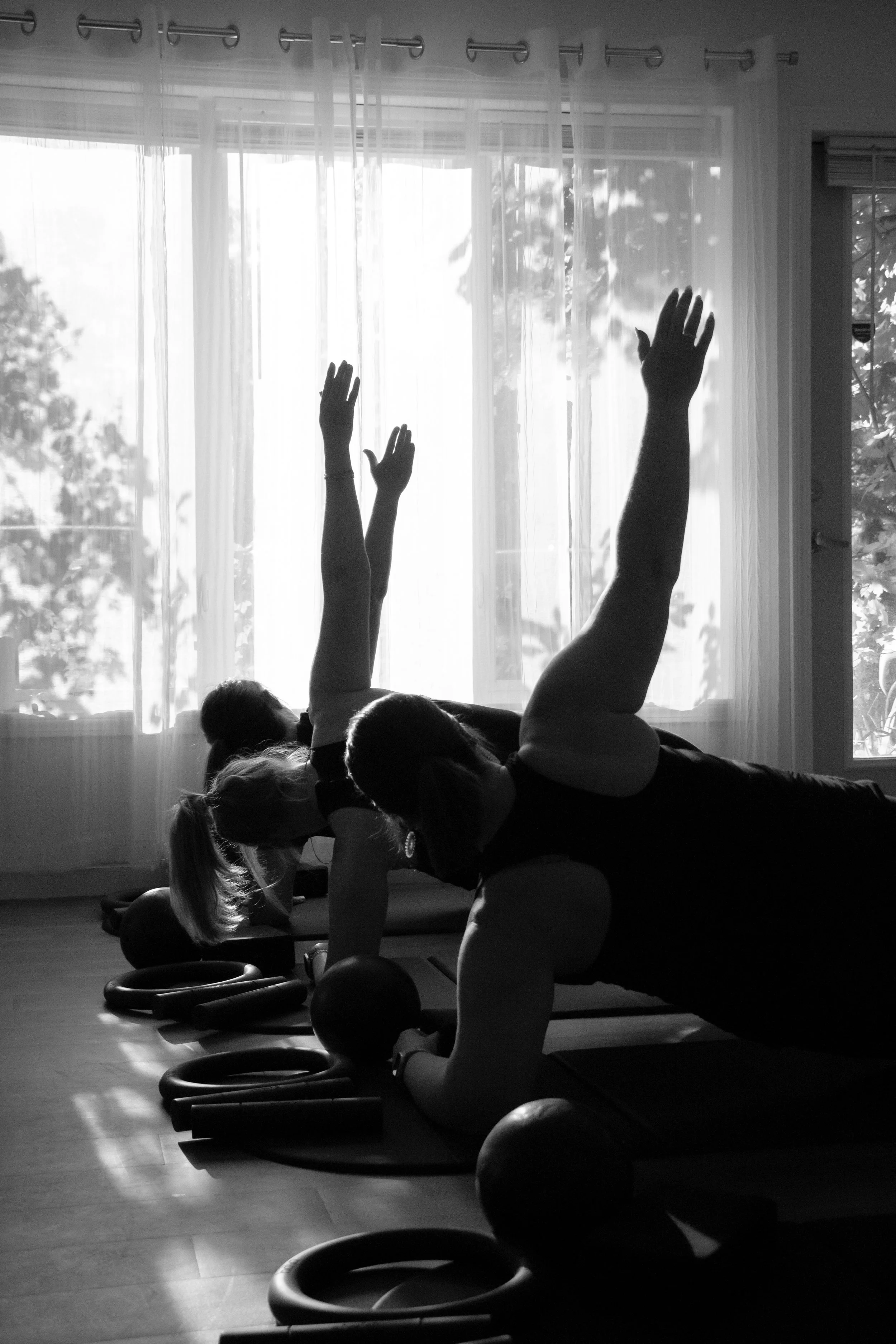 Two people doing yoga poses on mats in front of a large window with sheer curtains, with sunlight streaming in.