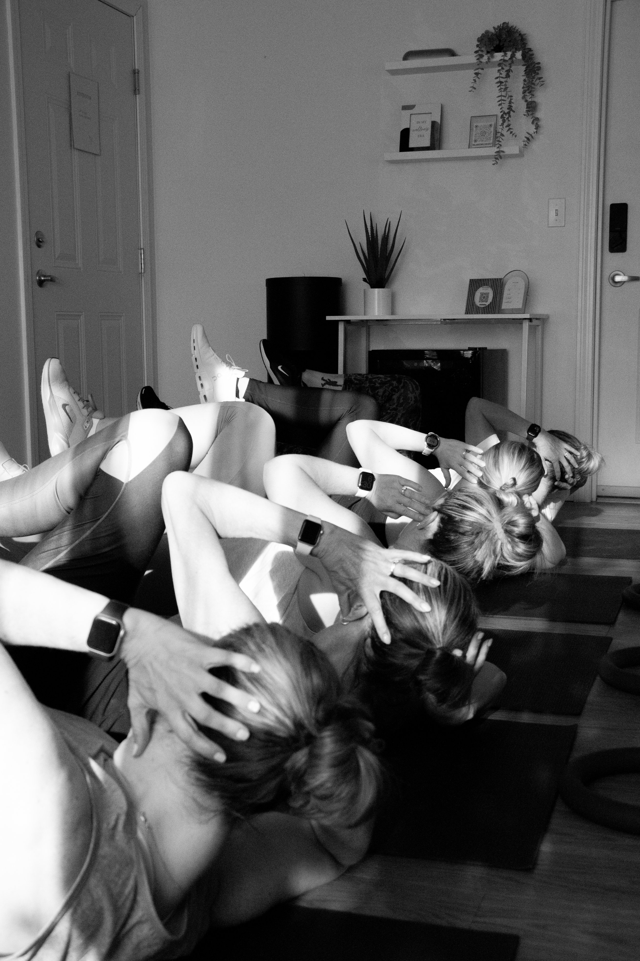 A group of women lying on yoga mats in a room, performing a yoga pose with their hands covering their ears and heads resting on the floor. They are wearing athletic clothing and smartwatches.