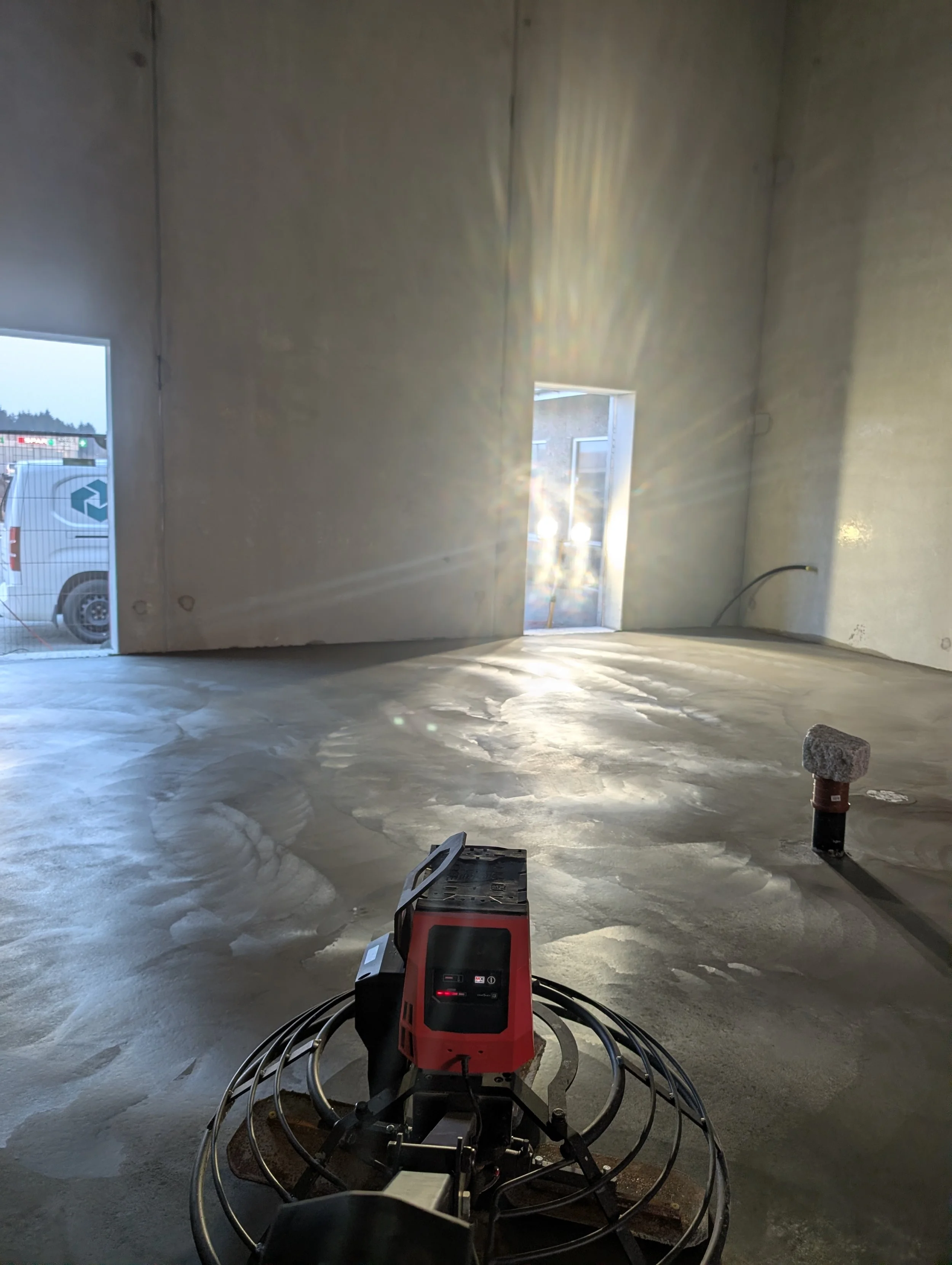 Innendørs byggeplass med betonggulv, to åpne vinduer, og elektrisk utstyr plassert på gulvet, klare for videre konstruksjon.