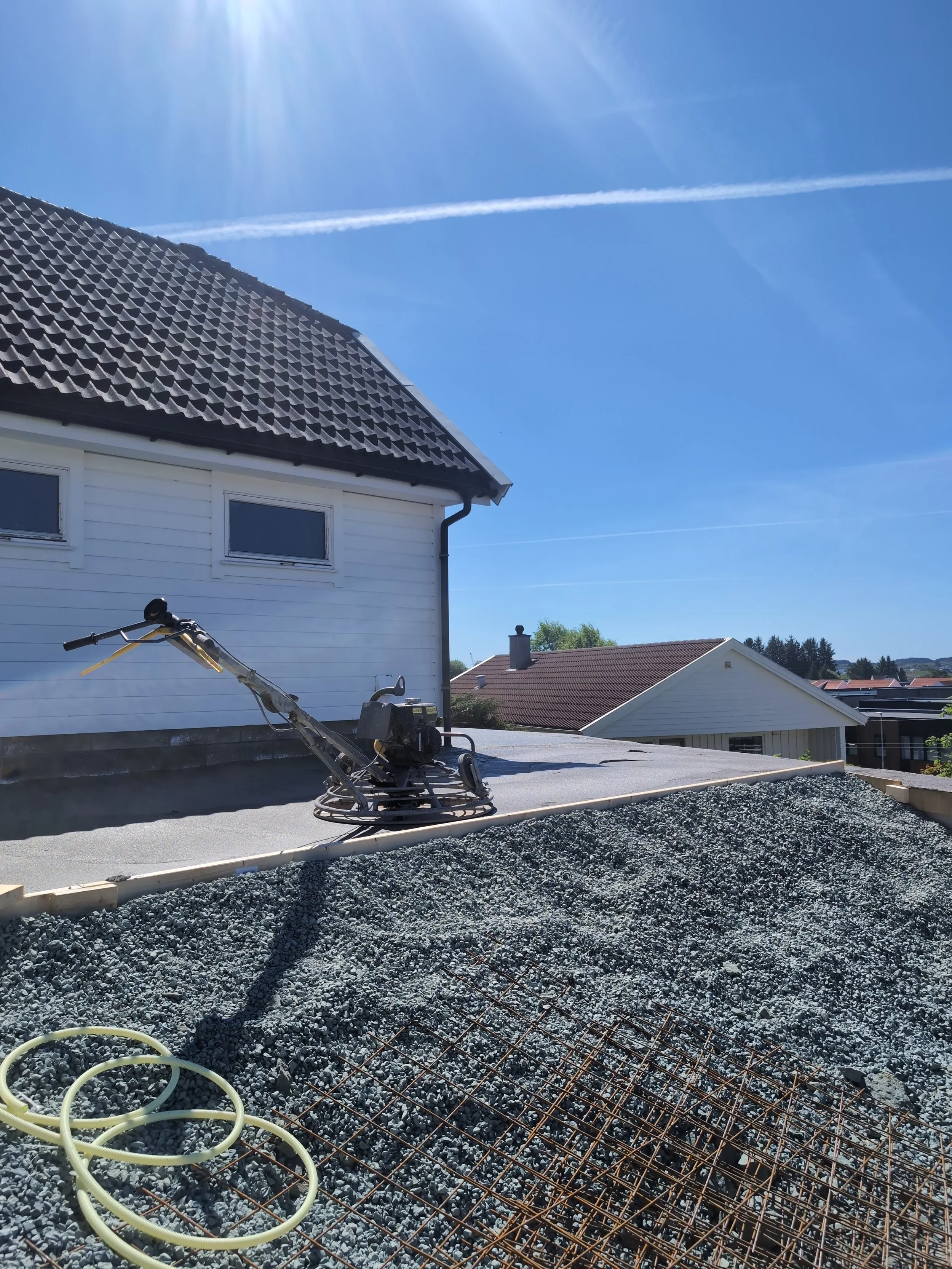 Støttemur under bygging med reir av grus og armeringsstål, og en maskin for betongarbeid i forgrunnen, på en solrik dag.