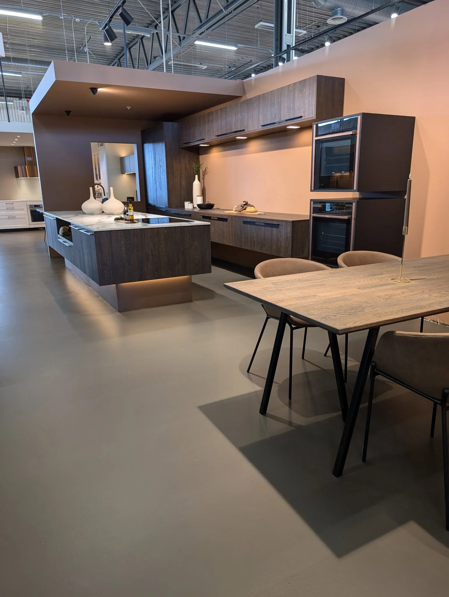 Moderne kjøkkenutstilling med mørk trepanel, hvit marmorøy, og beige vegg. Vaser og matvarer som brød og olje på kjøkkenøy. Spisebord med stoler i forgrunnen.