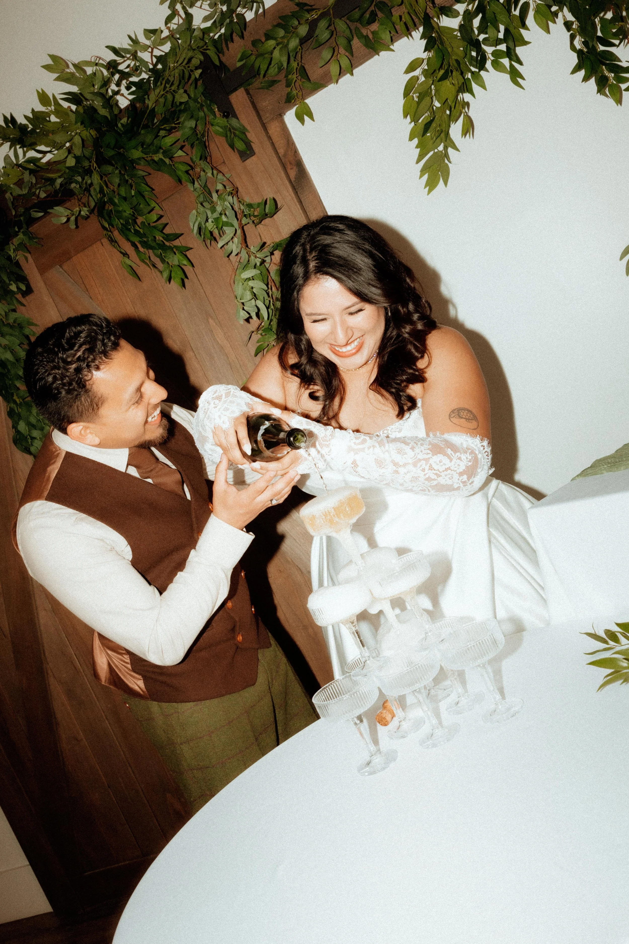 A bride and groom celebrating at a wedding reception, pouring champagne into a tower of glasses. They are smiling and looking at each other, with a white wall and green foliage in the background.