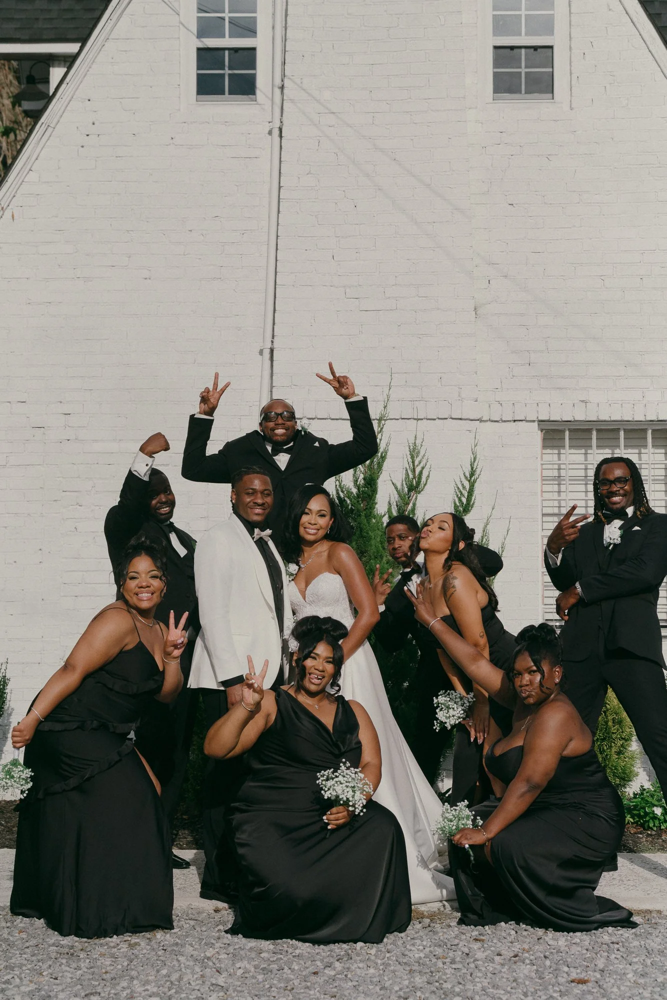 A group of people dressed in formal wedding attire posing outdoors in front of a white brick building. The bride wears a white gown, with others in black dresses or tuxedos, making peace signs and smiling.