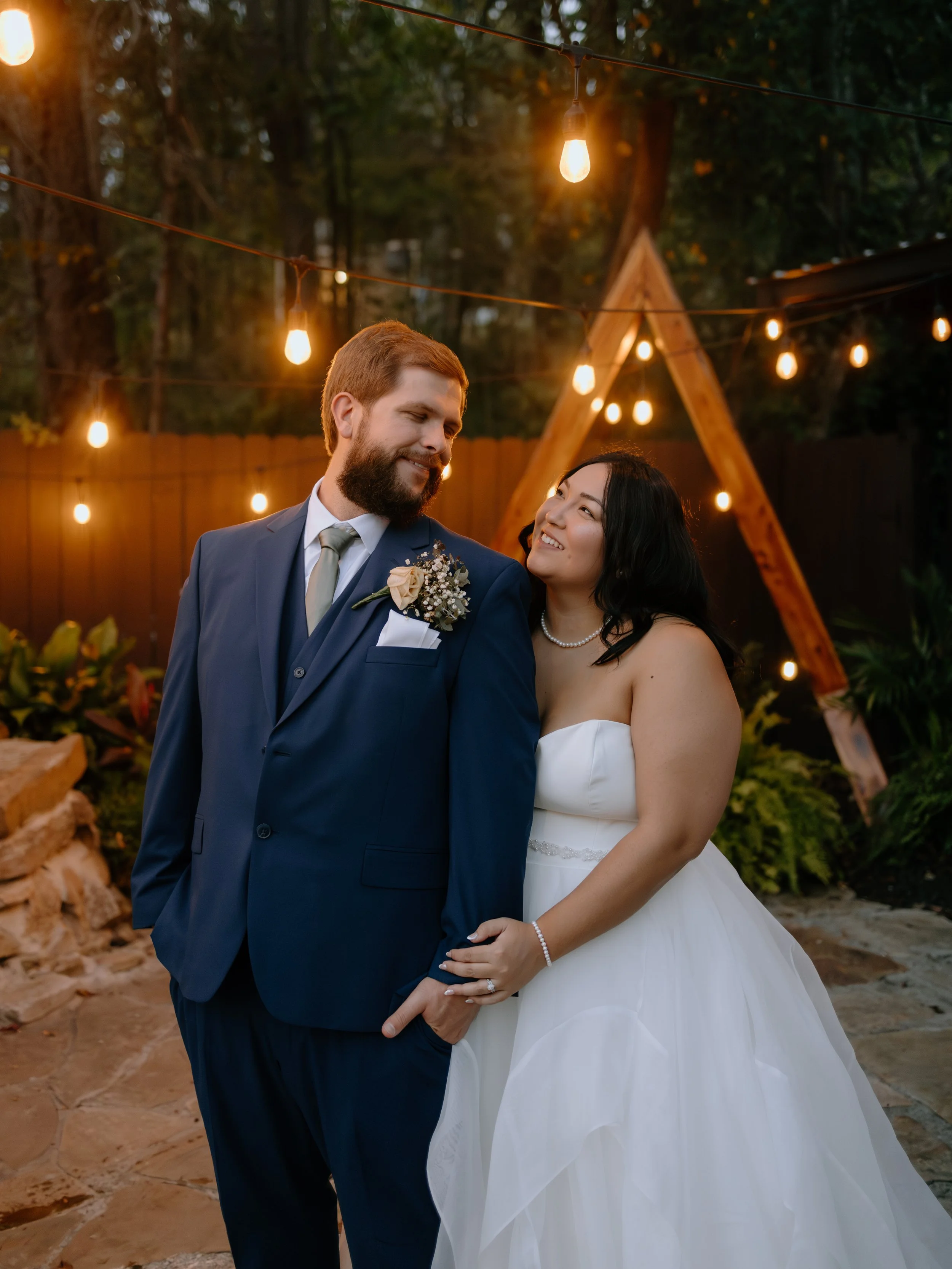 A wedding couple with the groom in a blue suit and the bride in a white dress, standing outdoors under warm string lights during the evening.