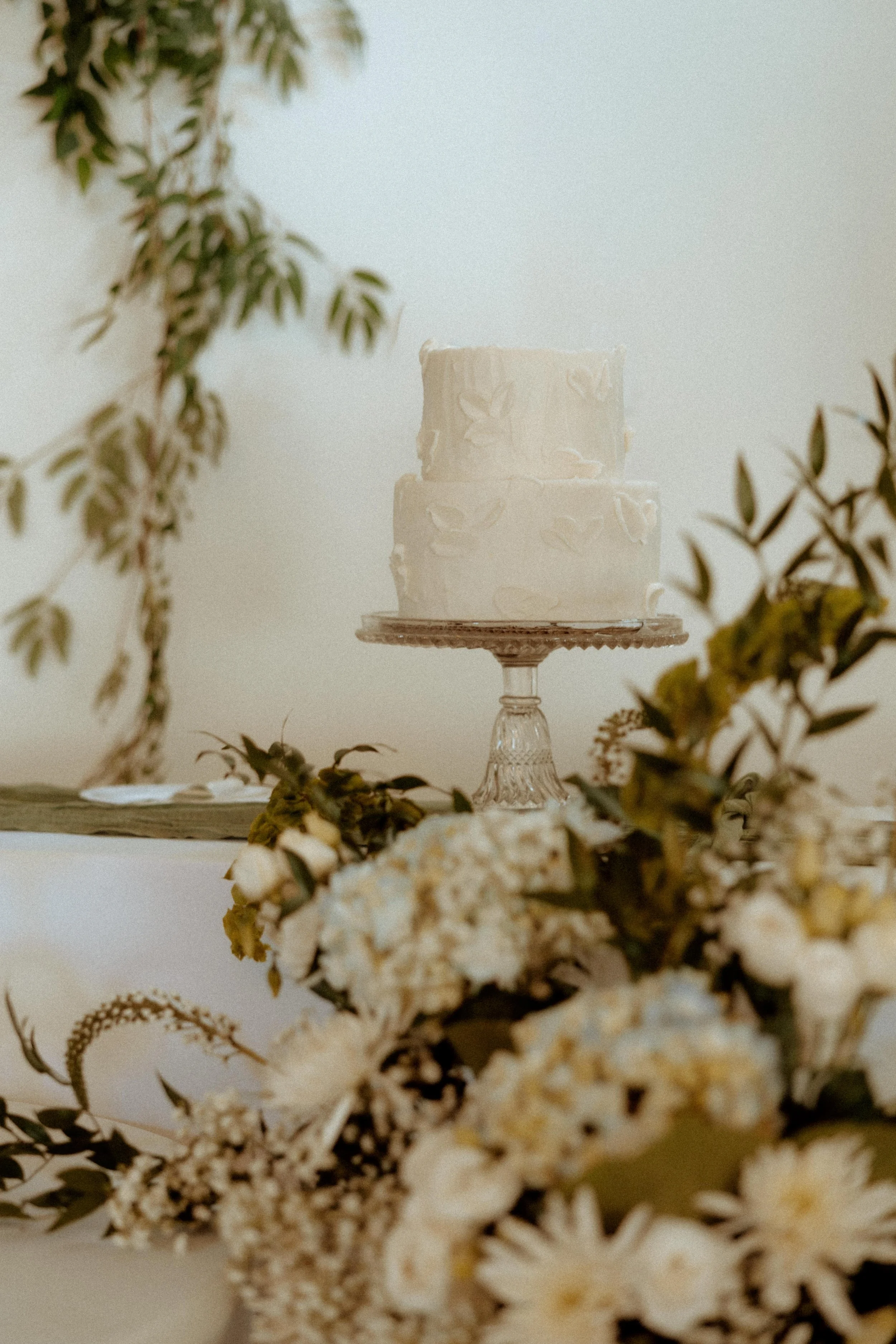 A white, two-tiered cake with a textured icing design, placed on a glass cake stand, surrounded by flowers and greenery for decoration.