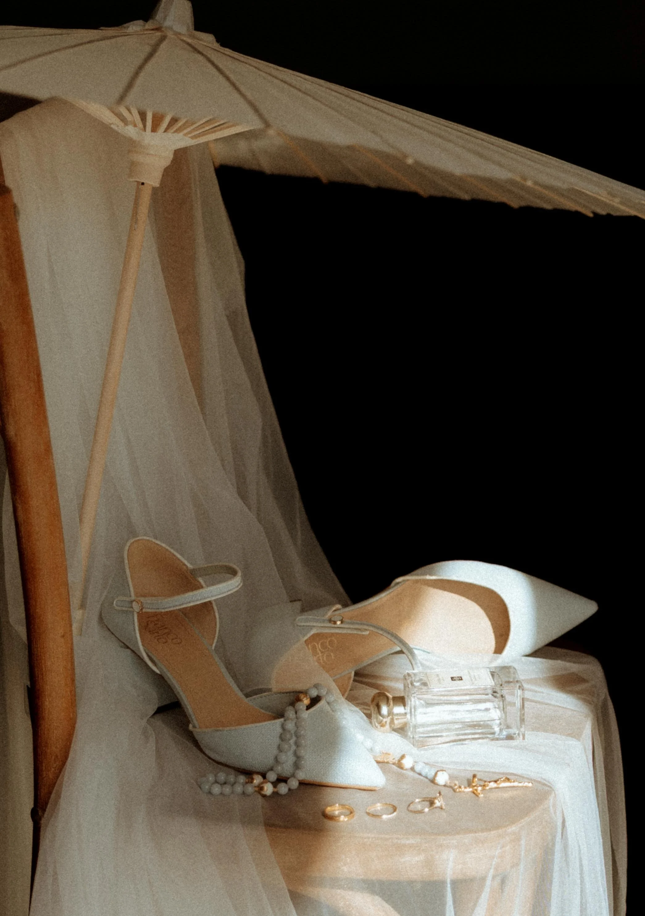 Display of white high-heeled shoes, pearl jewelry, and accessories on a table with a white cloth and a beige parasol.