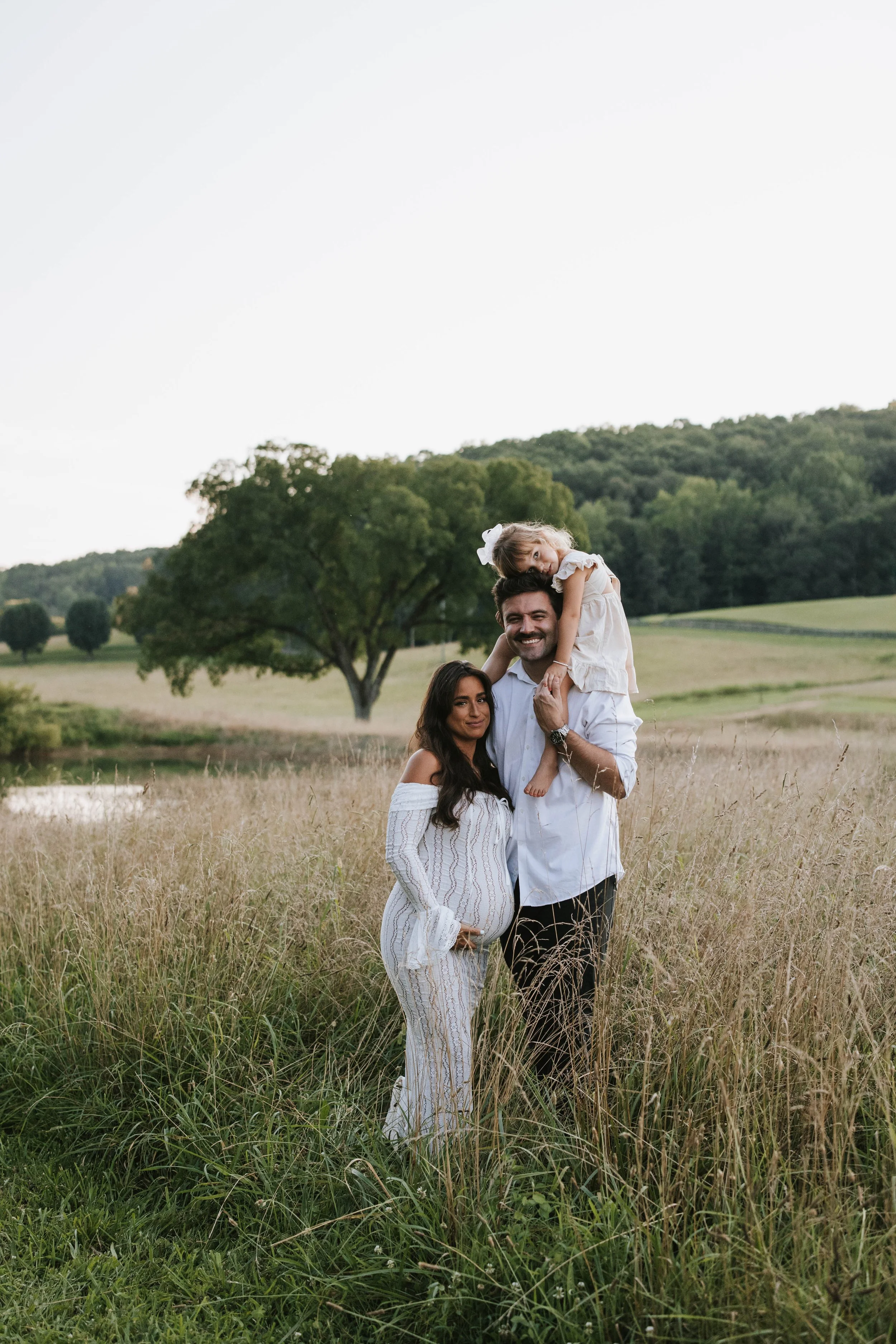 A family of three standing in tall grass in a rural outdoor setting, with trees and rolling hills in the background. The father is holding a young girl on his shoulders, and the mother is standing beside them, smiling.