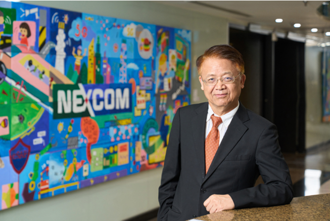 A man in a dark suit and orange tie standing in front of a colorful mural with the text 'NEXCOM' in an indoor setting.