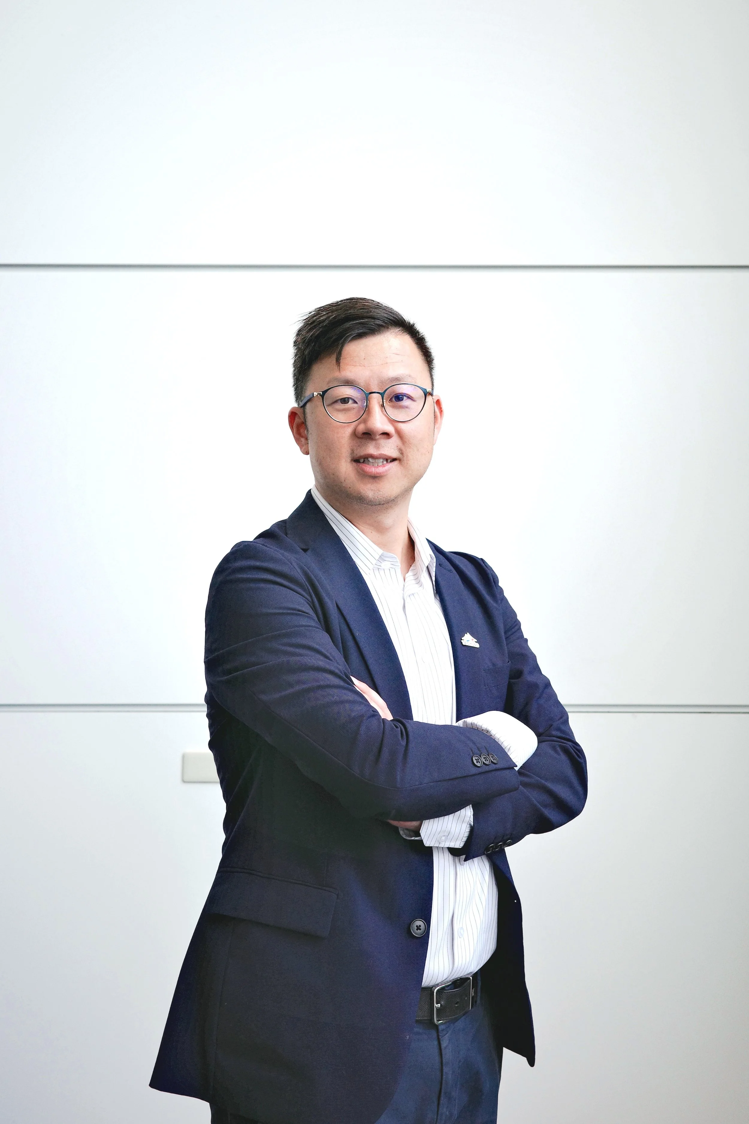 A professional man in a navy blue blazer, light striped dress shirt, and glasses stands with arms crossed in front of a plain white wall.