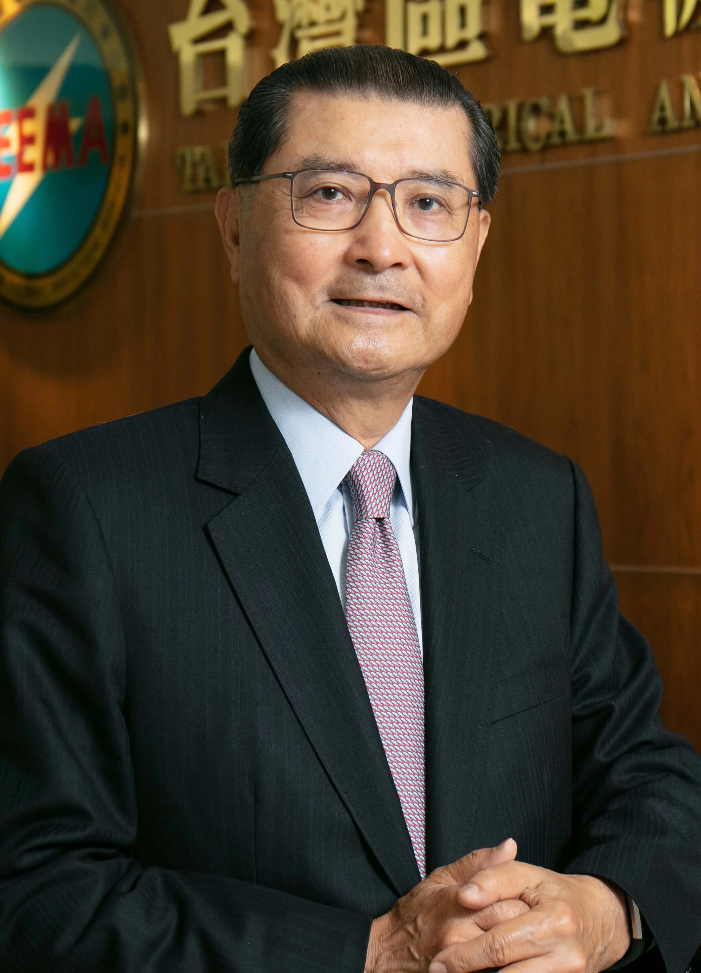 A middle-aged Asian man in a formal black suit, white shirt, and patterned tie, wearing glasses, standing with hands clasped, in front of a wooden background with a wall sign and a colorful emblem.