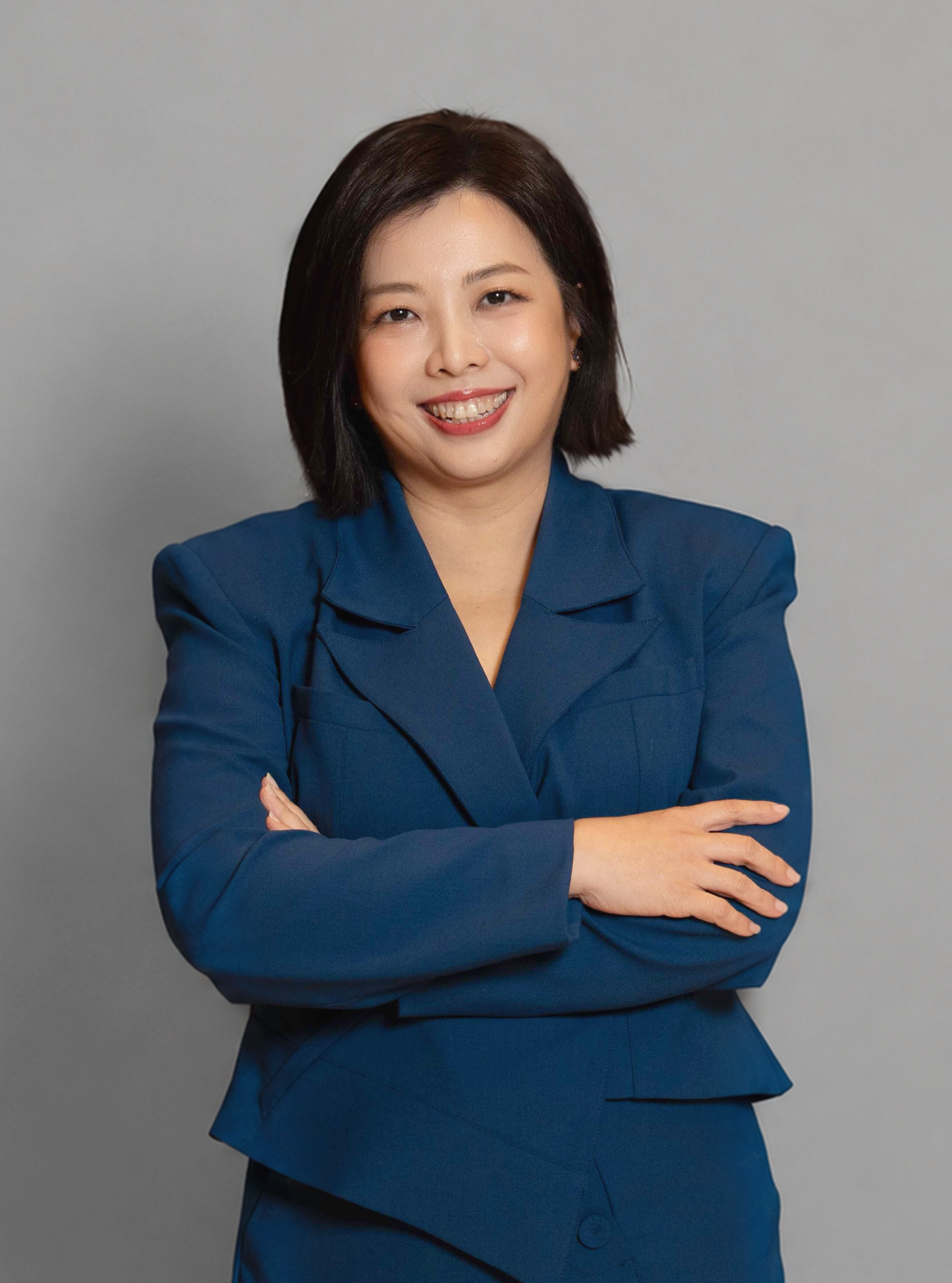 A woman with straight, shoulder-length dark hair, smiling and winking, wearing a navy blue blazer and skirt, with arms crossed in front of a plain gray background.