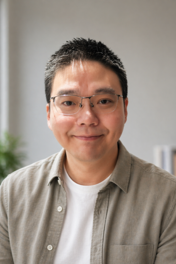 Portrait of a man with short black hair, glasses, wearing a beige button-up shirt over a white t-shirt, smiling.