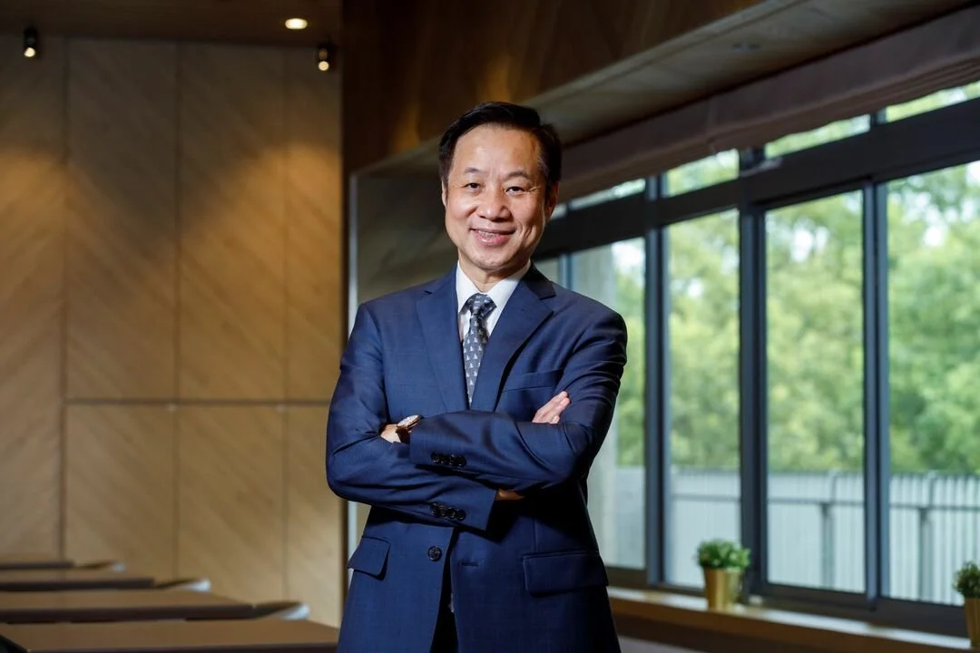 A man in a blue suit smiling with arms crossed in an office with large windows and green trees outside.