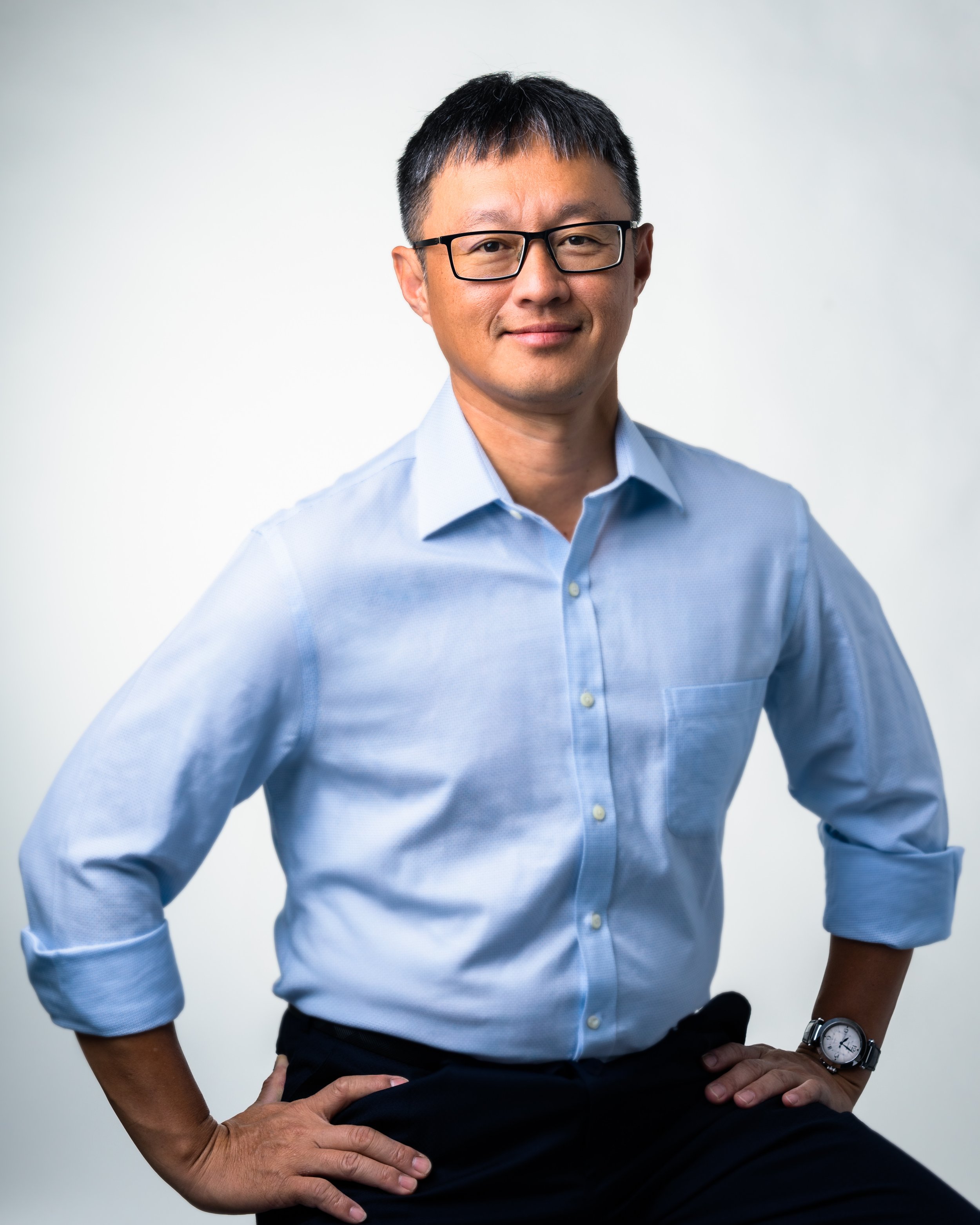 Close-up image of a middle-aged man with black hair, wearing glasses, a light blue dress shirt, black pants, and a wristwatch, standing against a plain light background with his hands on his hips.