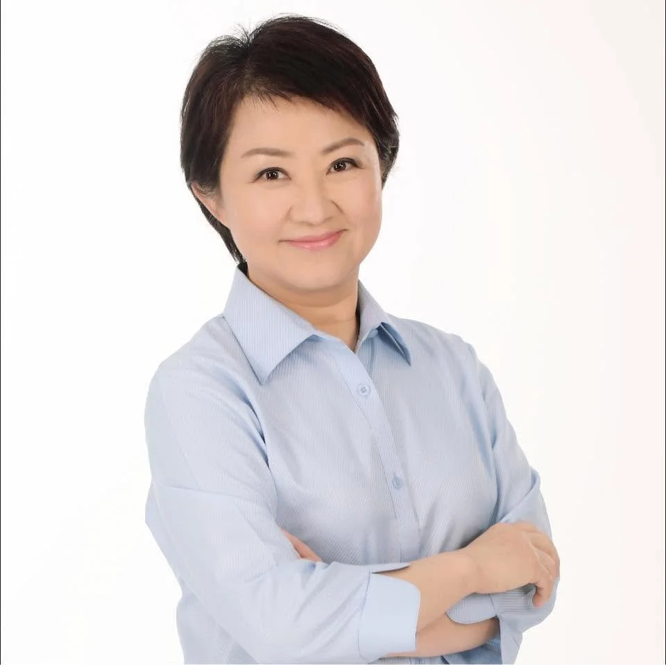 A woman with short dark hair wearing a light blue button-up shirt, smiling with her arms crossed in front of a plain white background.