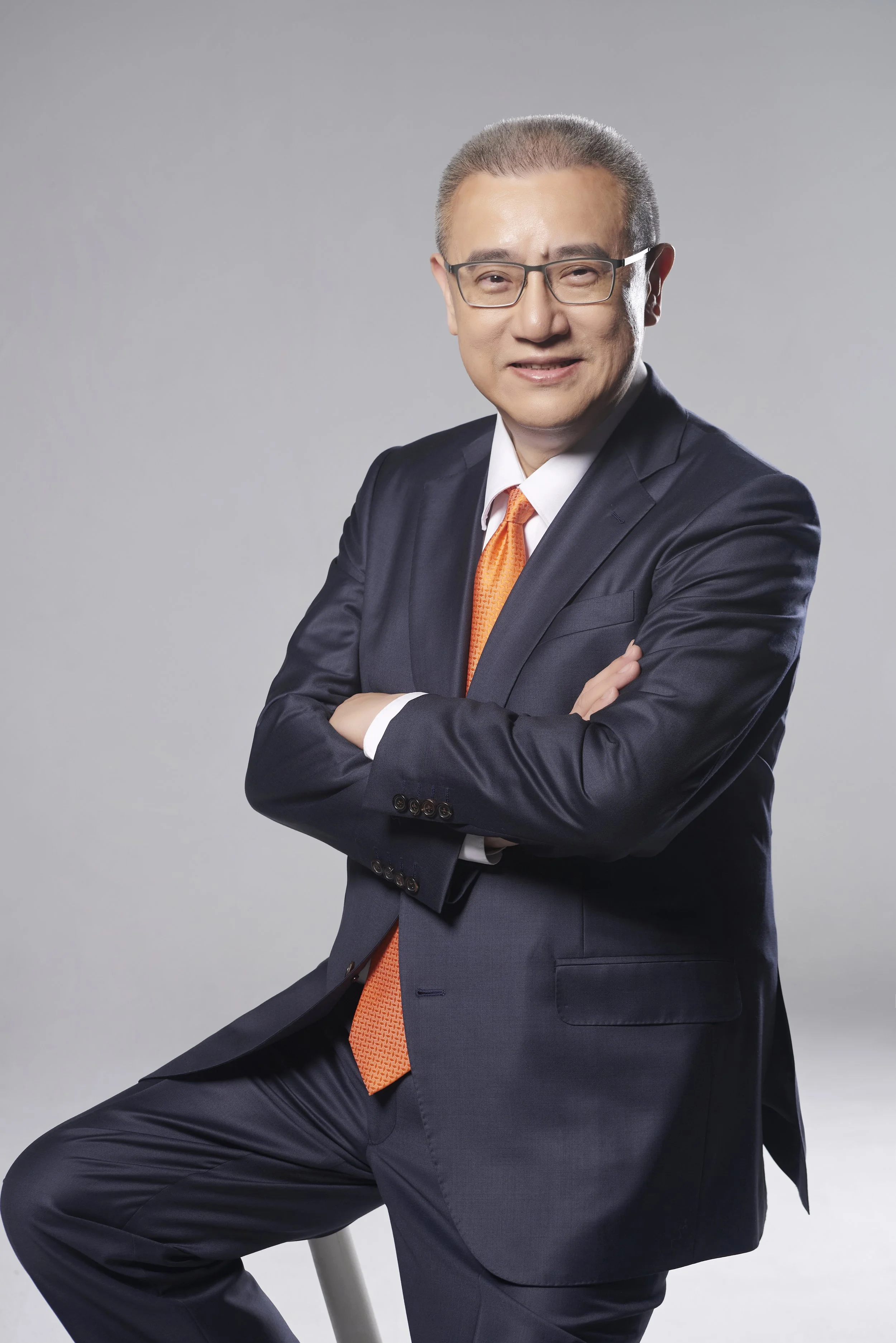 A professional man in a dark suit with an orange tie, wearing glasses, sitting on a stool with arms crossed, smiling at the camera, against a neutral gray background.