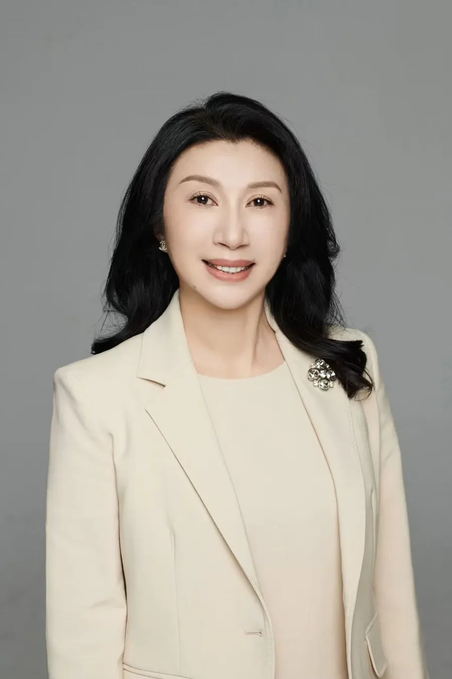 A professional woman with long black hair smiling, wearing a cream-colored blazer with a brooch, earrings, and a cream top, against a plain grey background.