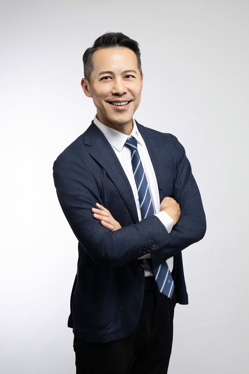A smiling man in a business suit with arms crossed, wearing a navy blazer, white shirt, and striped tie, standing against a plain white background.
