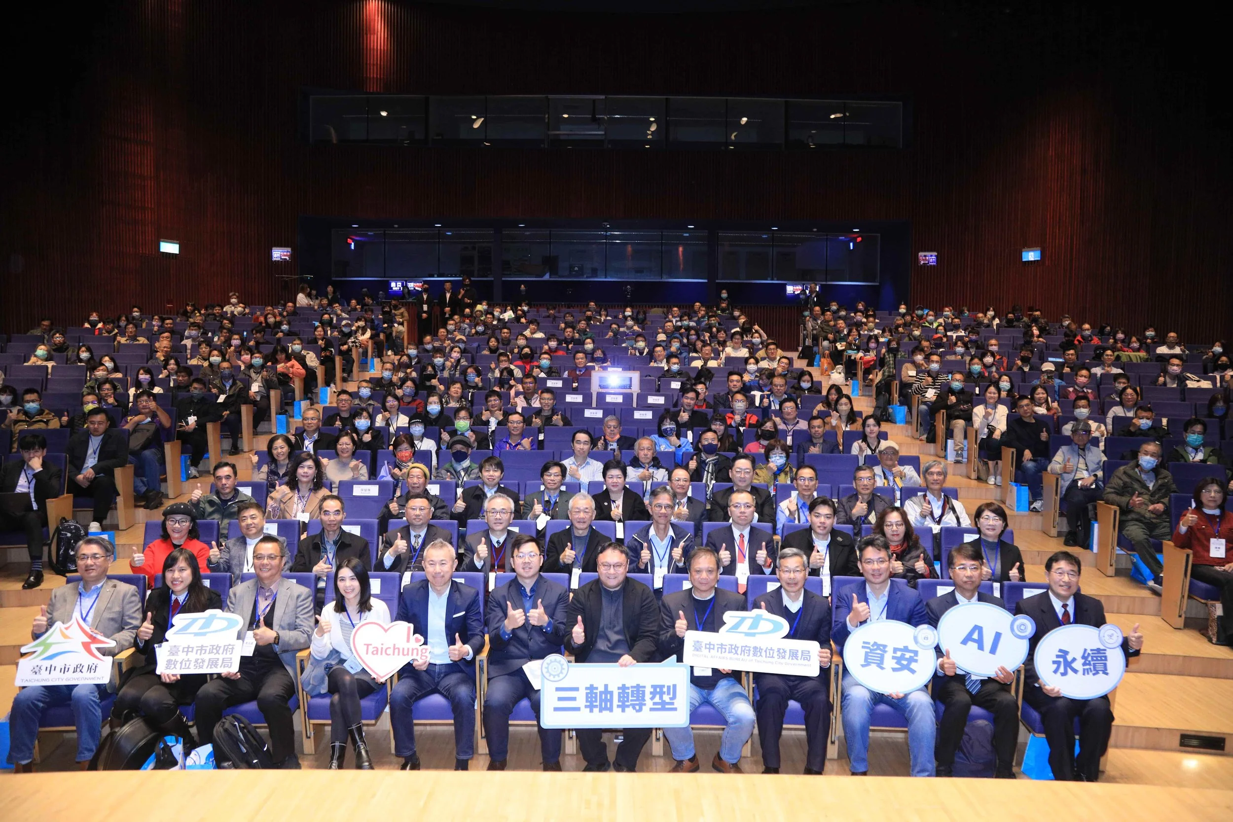 Group of people attending a conference or seminar in a large auditorium, many wearing face masks, with some holding signs and giving thumbs up.