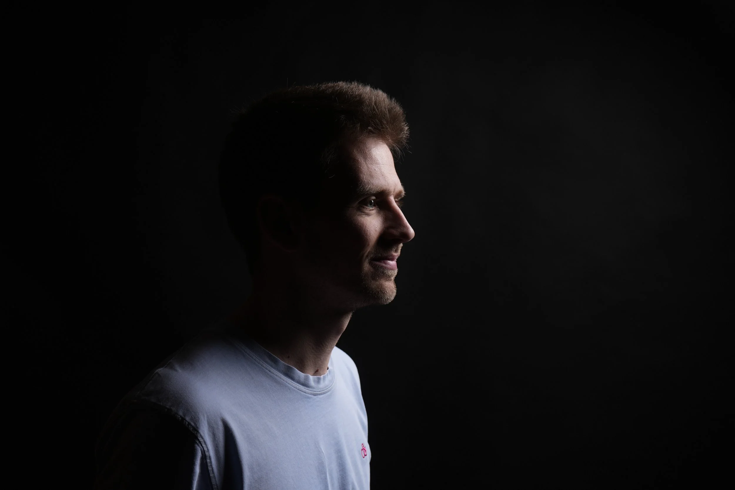 Profile of a young man with short brown hair and a beard, wearing a light gray shirt, against a black background.