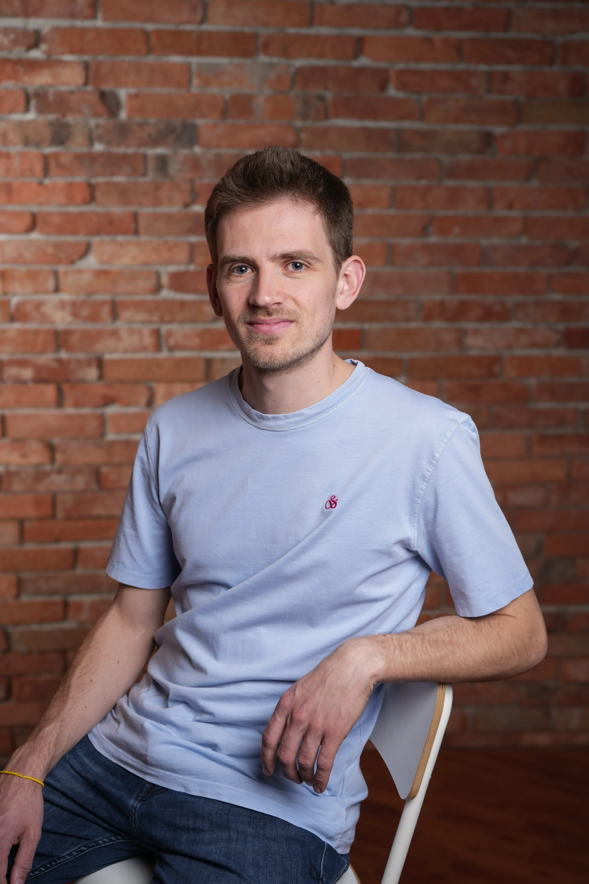 A young man with short brown hair and light skin sitting on a chair in front of a brick wall. He is wearing a light blue t-shirt and dark jeans, and has a relaxed pose with his right arm resting on his thigh and his left arm on the armrest. He has a slight smile and is looking at the camera.