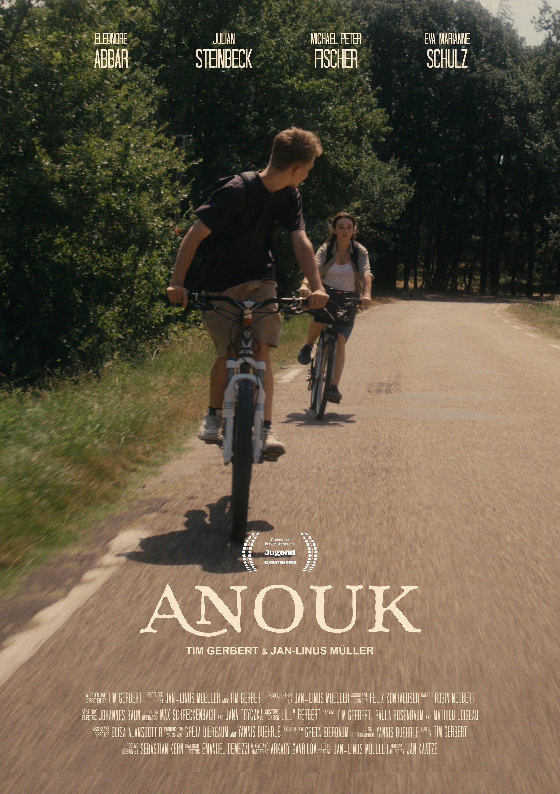 Two young people riding bicycles on a dirt road surrounded by trees, with movie credits and title "Anouk" overlaid on the image.