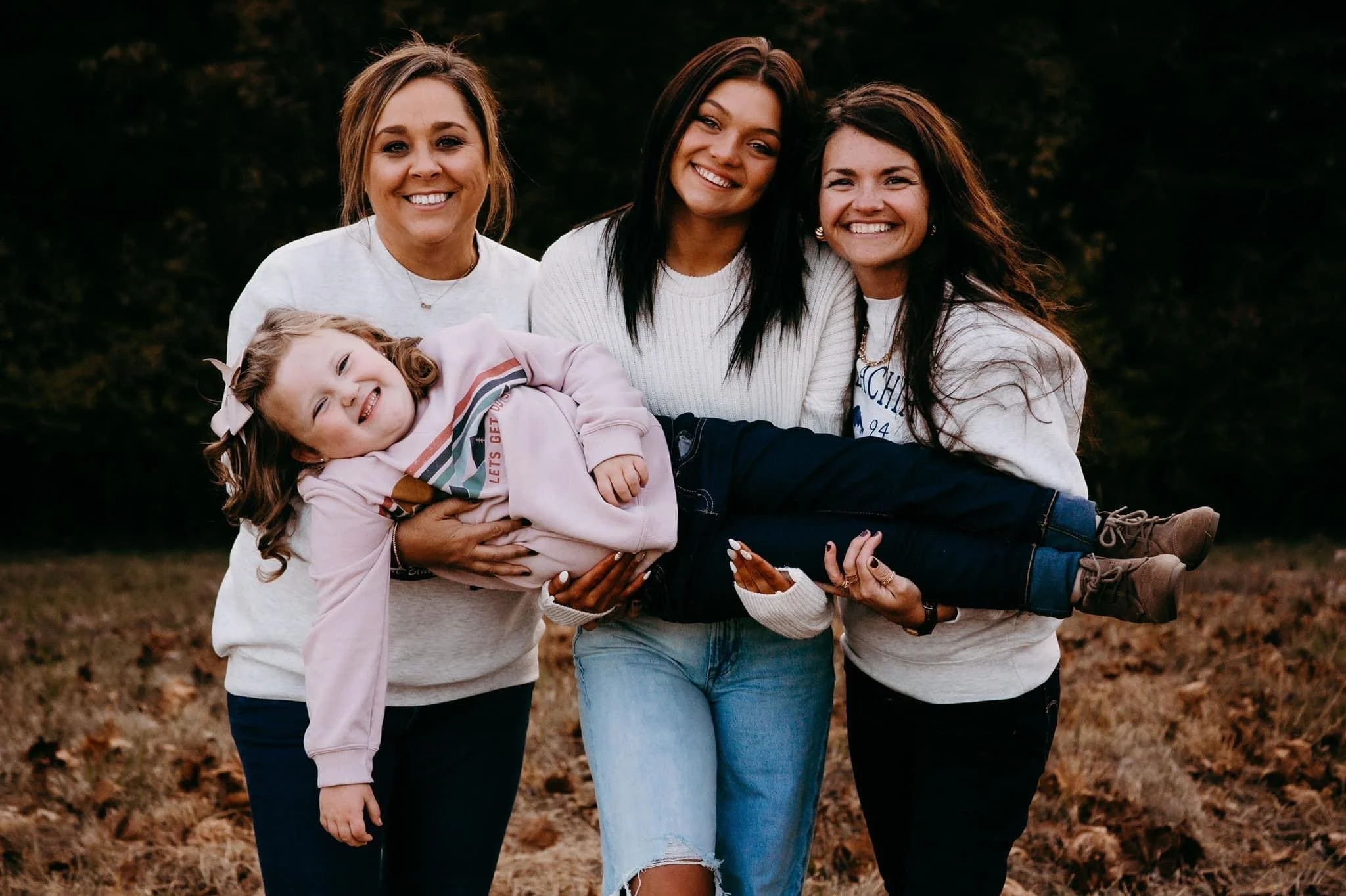 Four women and a young girl outdoors, standing on fallen leaves, smiling and holding each other, with a leafy background.
