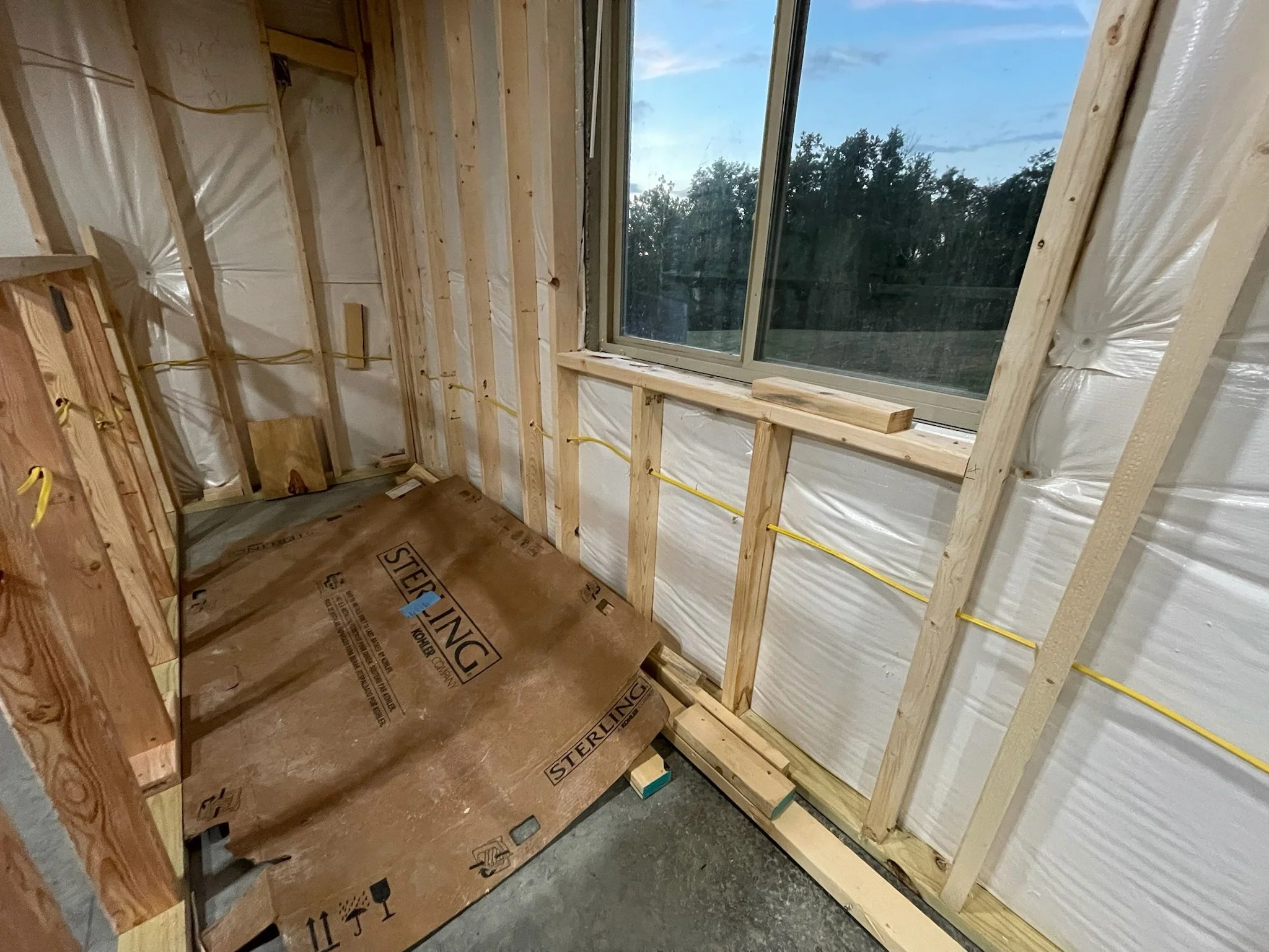 Interior of a building under construction with visible wooden framing, a large window showing trees outside, and a cardboard box on the floor.