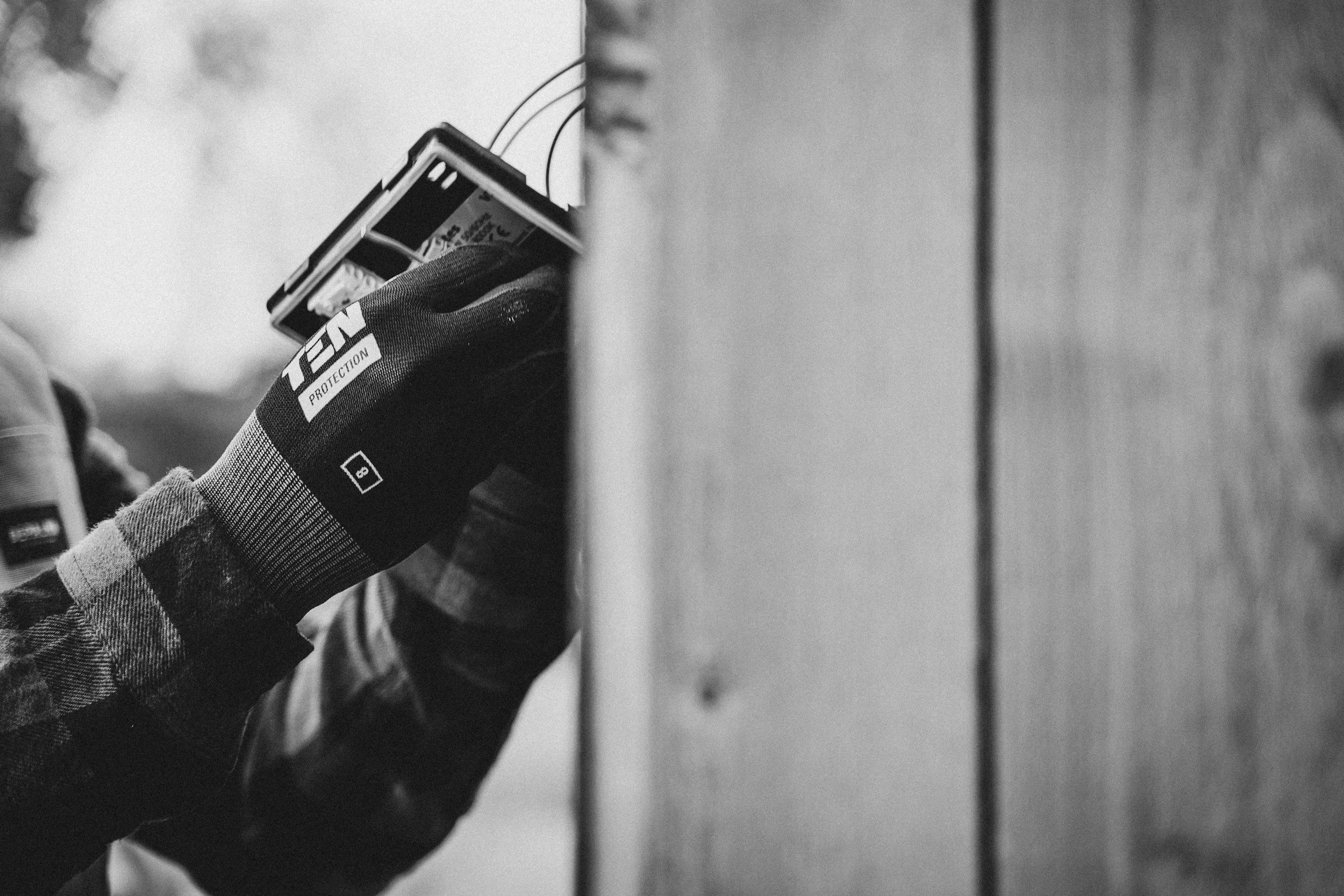 A person wearing a checkered glove holding a small electronic device, possibly a battery or a card, with wires attached, near a smooth vertical surface.