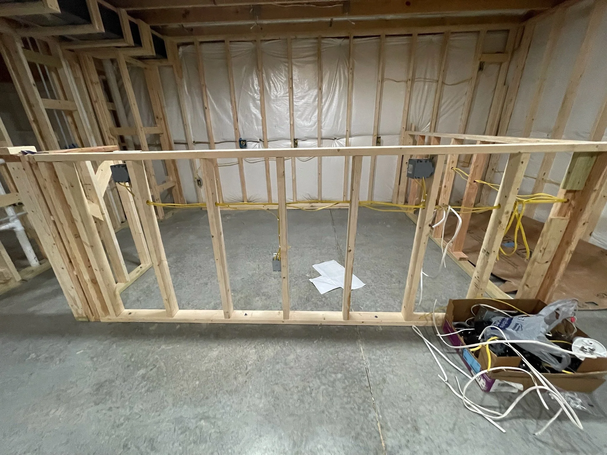 Interior view of a house under construction with exposed wooden framing, electrical wires, and construction materials on the floor.