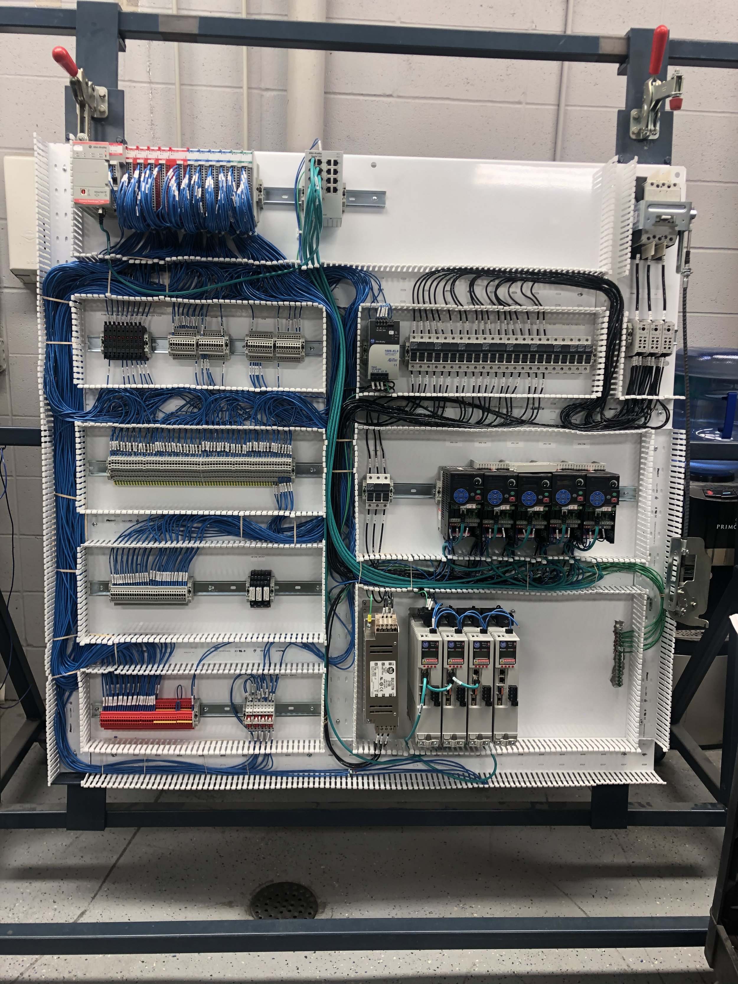 Electrical control panel with various wires, relays, and circuit boards, mounted on a white backboard within a metal frame.