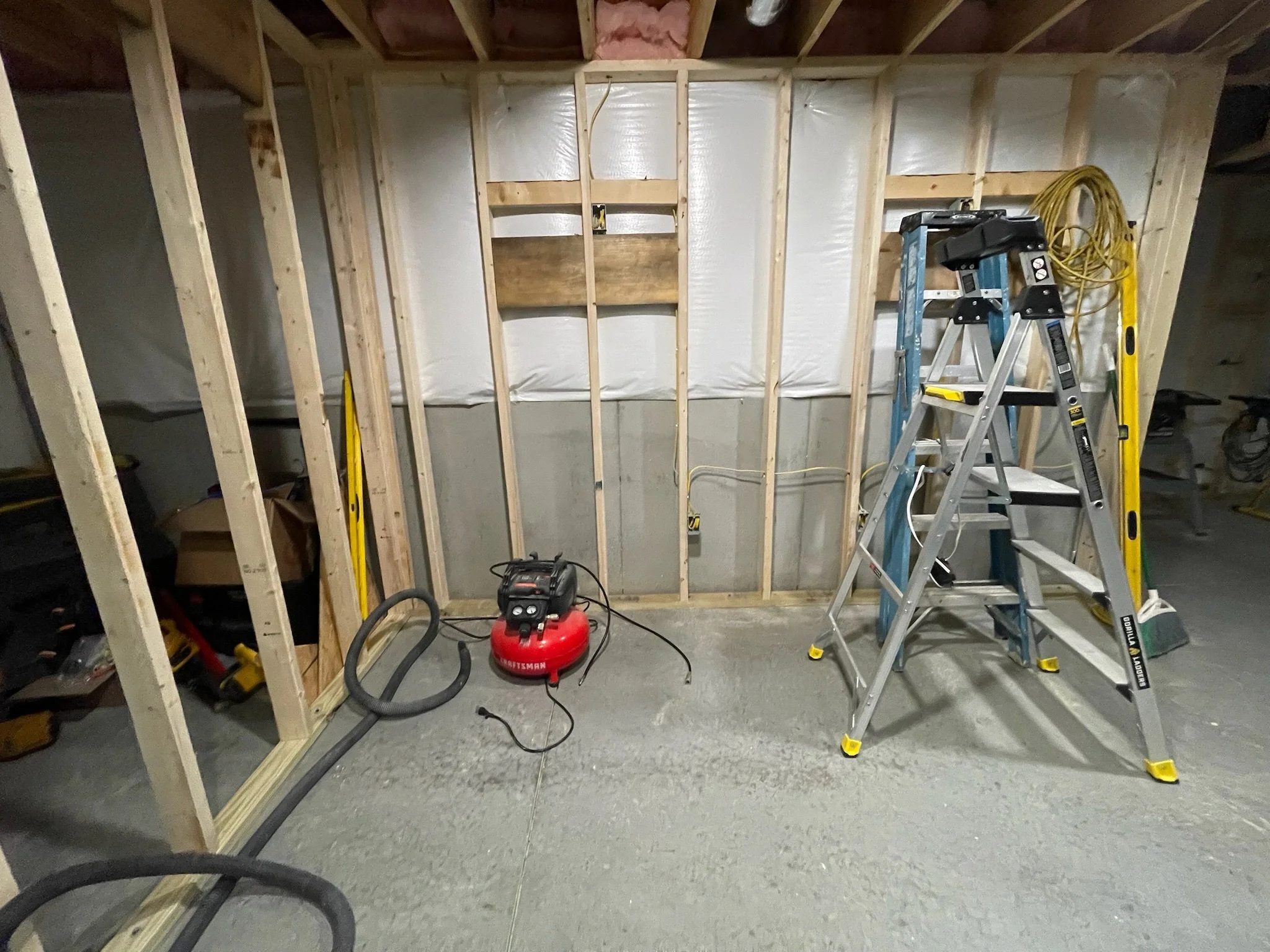 Unfinished wall framing with wooden studs, insulation, ladder, and tools in a basement or construction site.