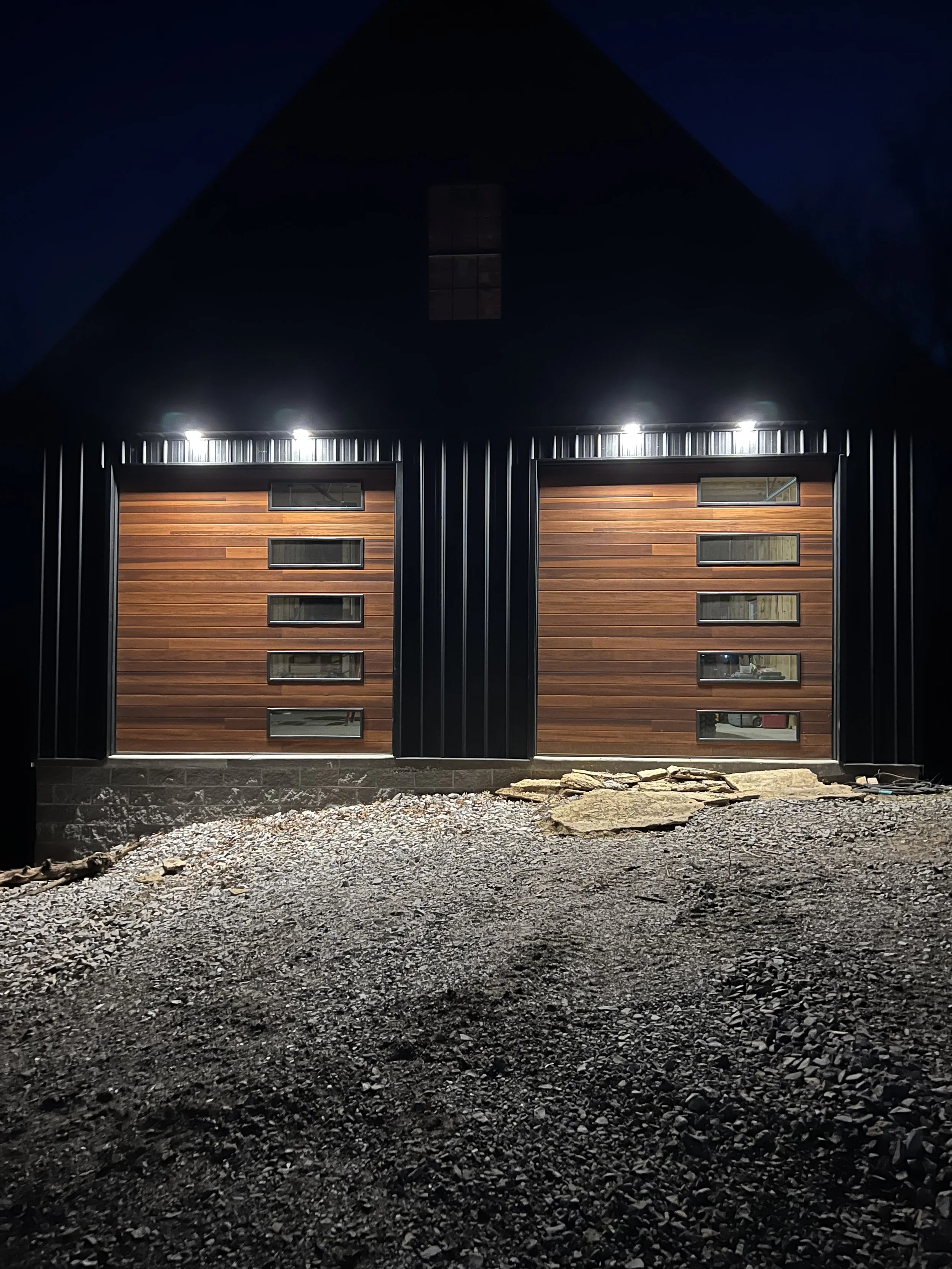 A modern house with garage doors featuring wooden panels, illuminated from above, situated on a gravel lot at night.