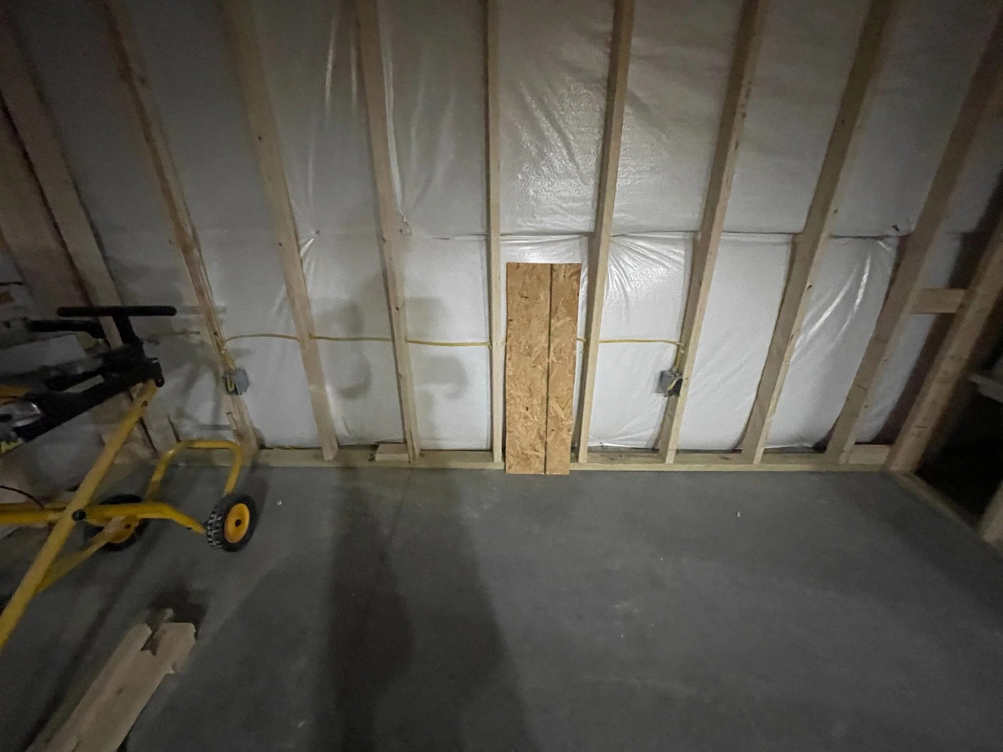 The image shows an unfinished room's ceiling with wooden beams, insulation covered with plastic sheeting, and electrical wiring. There are construction tools and equipment, such as a yellow hand truck, visible in the room.