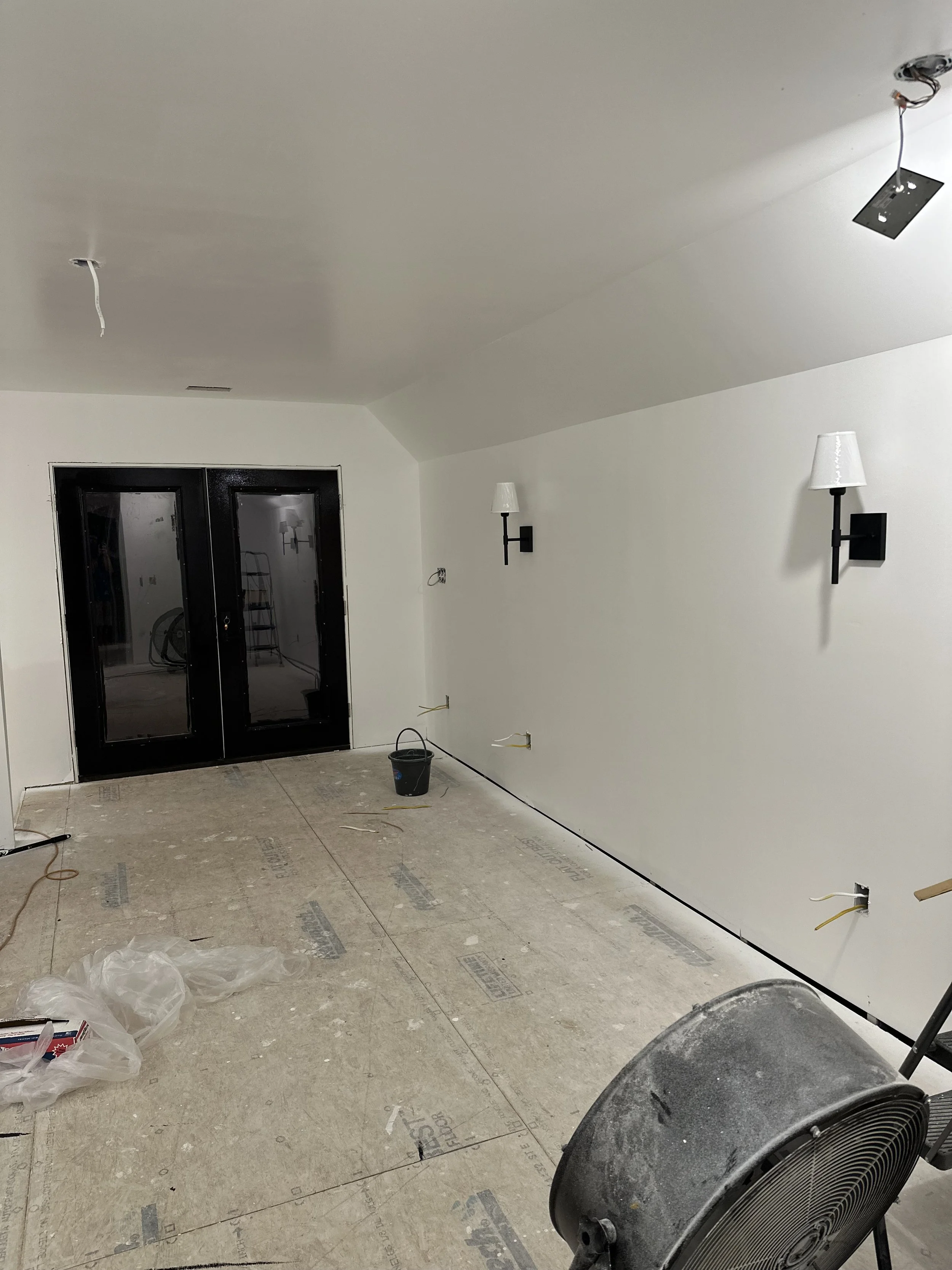 Interior room under construction with white walls, black double doors, unfinished flooring, and wall-mounted light fixtures. Construction tools and materials are on the floor, including a plastic bag, wire, and a small bucket. An oscillating fan is i