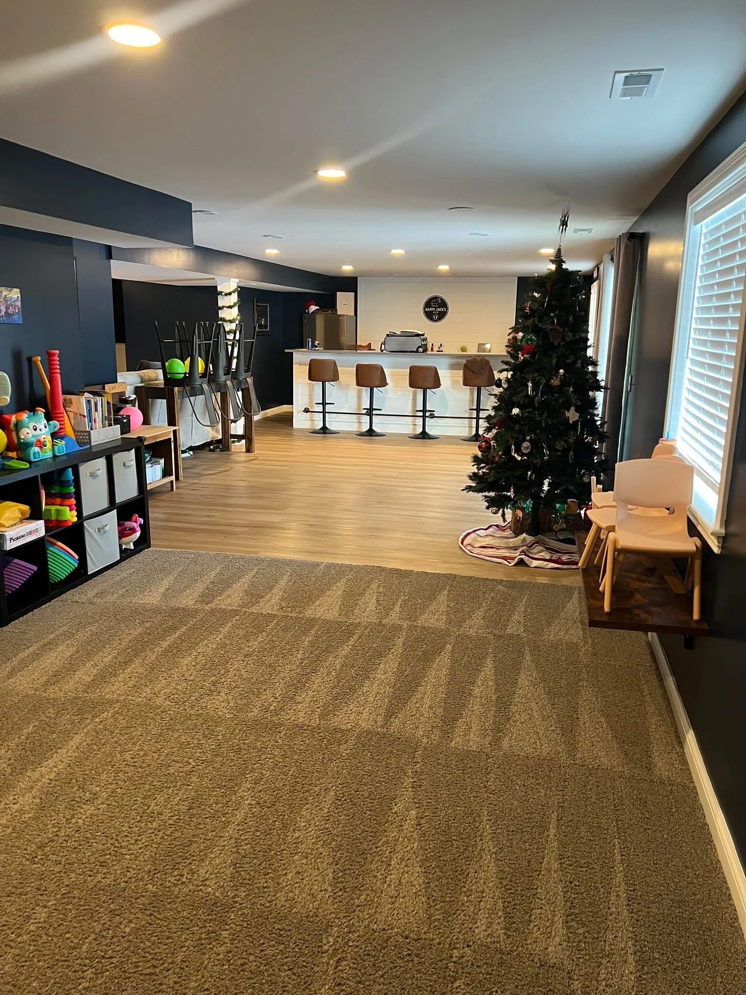 A living room decorated for Christmas with a Christmas tree, chairs, and toys, and a bar area in the background.