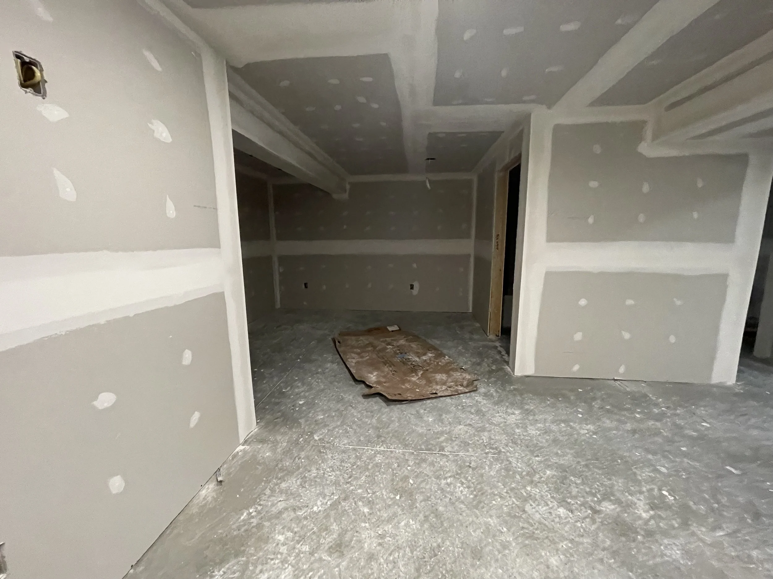 An unfinished room under construction with drywall walls and ceiling with seams and screw holes, a concrete floor with a protective sheet, and a wooden board on the floor.
