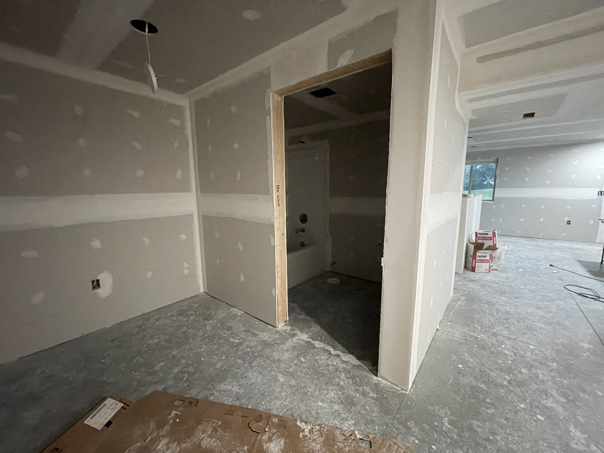 Interior of a house under construction with drywall installed on walls and ceiling, unfinished floor, and a doorway opening to another room, with construction tools and materials visible.