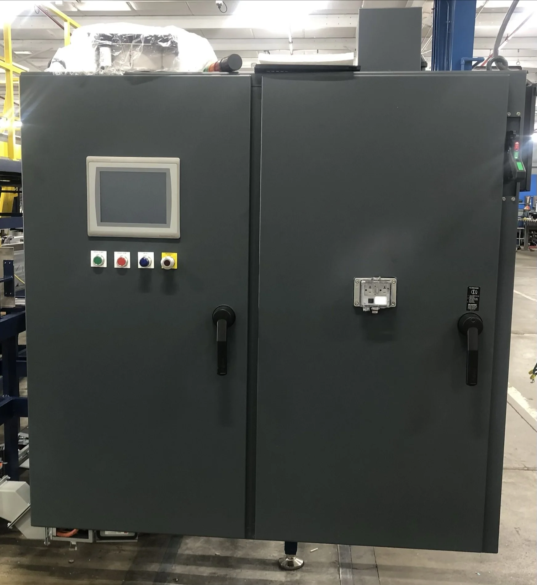 Industrial machine with a grey control panel featuring a touchscreen display and four colored buttons, located in a warehouse with metal shelving and platform in the background.