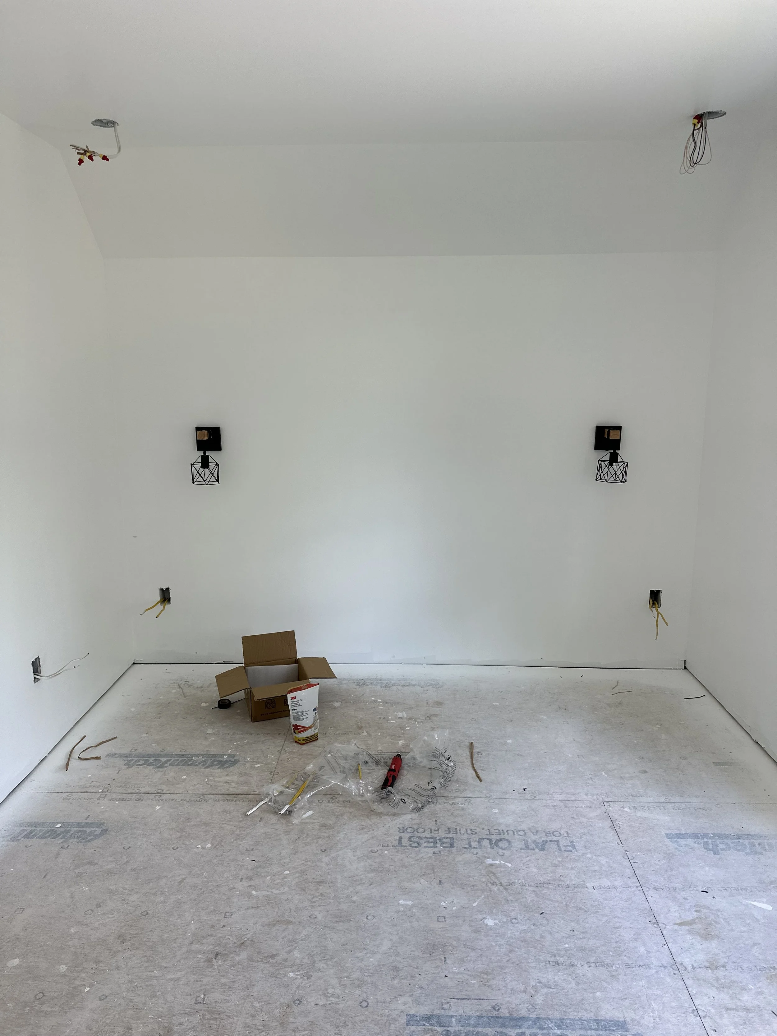 Empty room under construction with wires hanging from the ceiling, light fixtures not yet installed, a cardboard box and tools on the floor, and a protective sheet covering the floor.