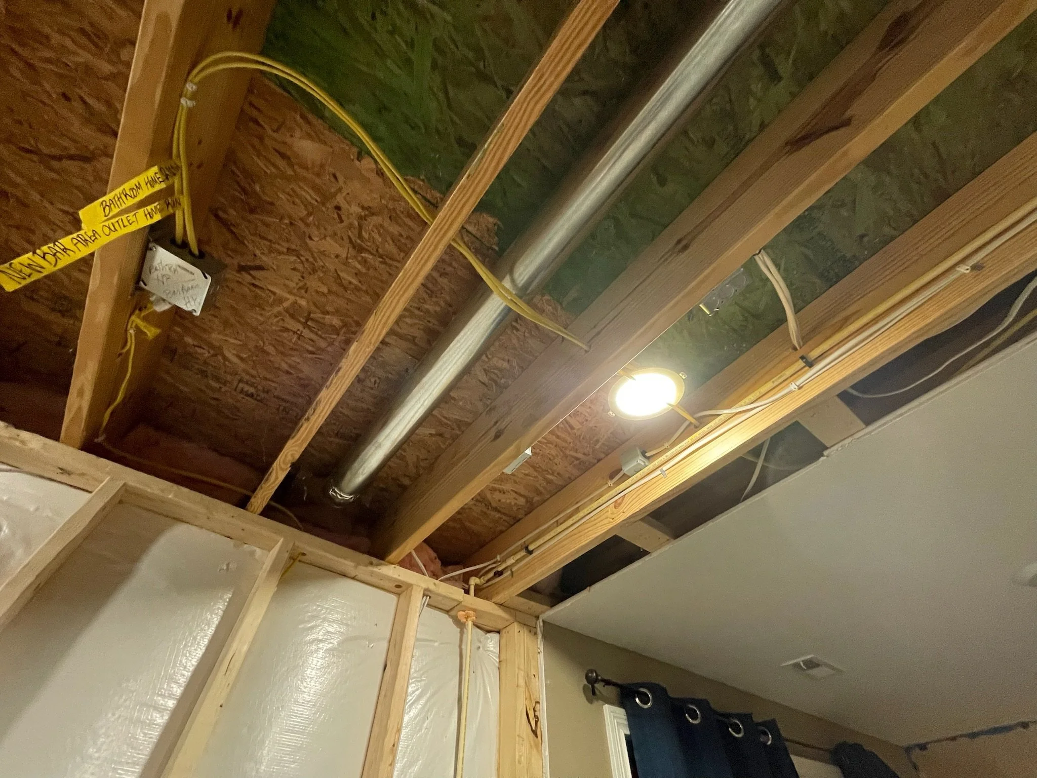 Ceiling framing with wooden beams, insulation, metal ductwork, electrical wires, and a light fixture in a basement or attic space.