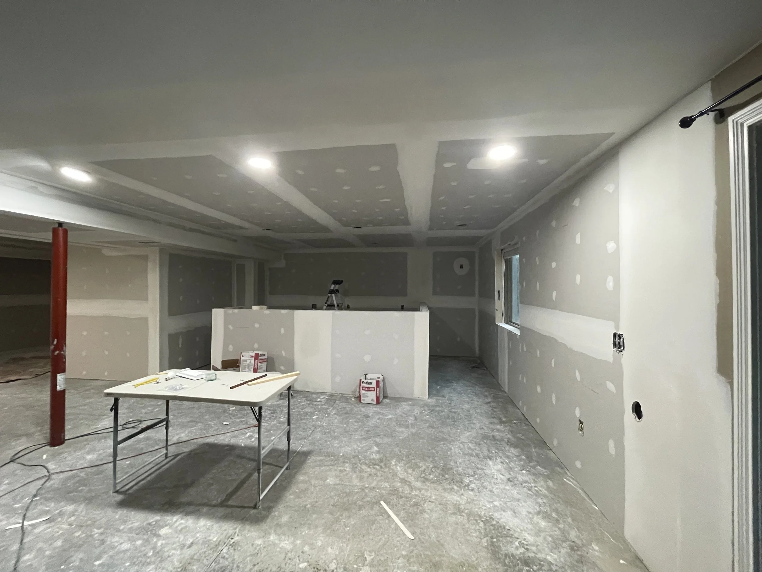 A room under construction with taped drywall, a ladder, a folding table with construction tools, a red pole, and boxes on the floor.