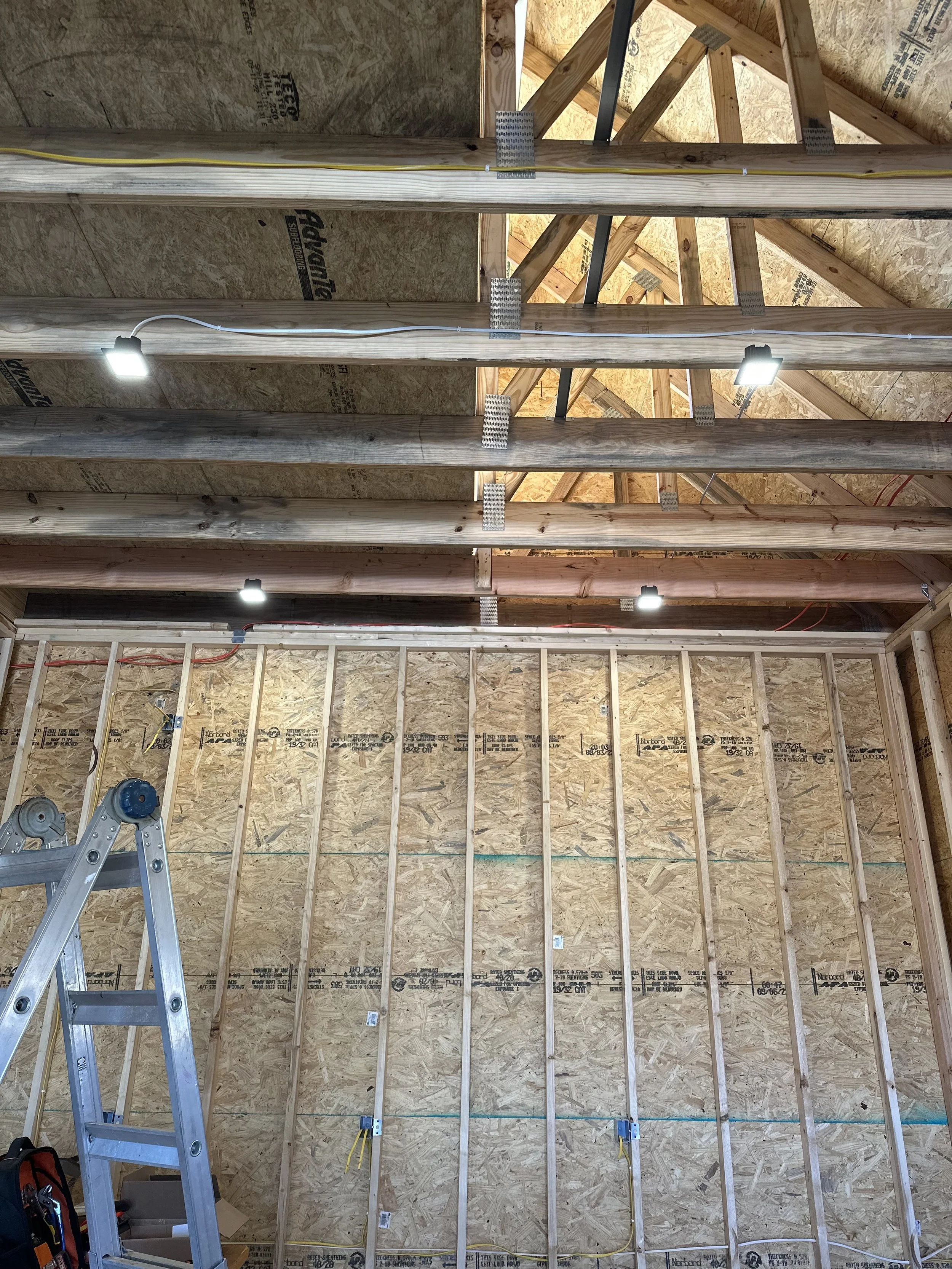 Interior view of a construction site showing wooden framing, exposed OSB wall sheathing, electrical wiring, metal brackets, overhead lighting, and a ladder, indicating ongoing building process.