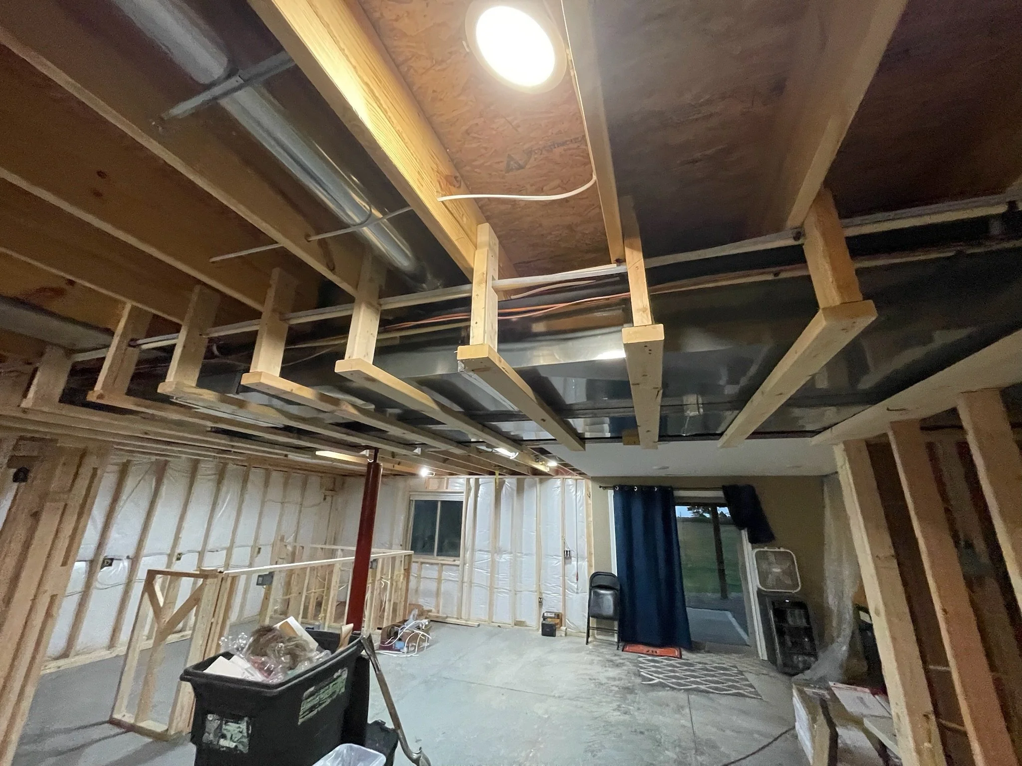 Interior of a house under construction with exposed wooden framing and HVAC ductwork, a ladder, a black storage bin, a chair, and construction materials on the floor.