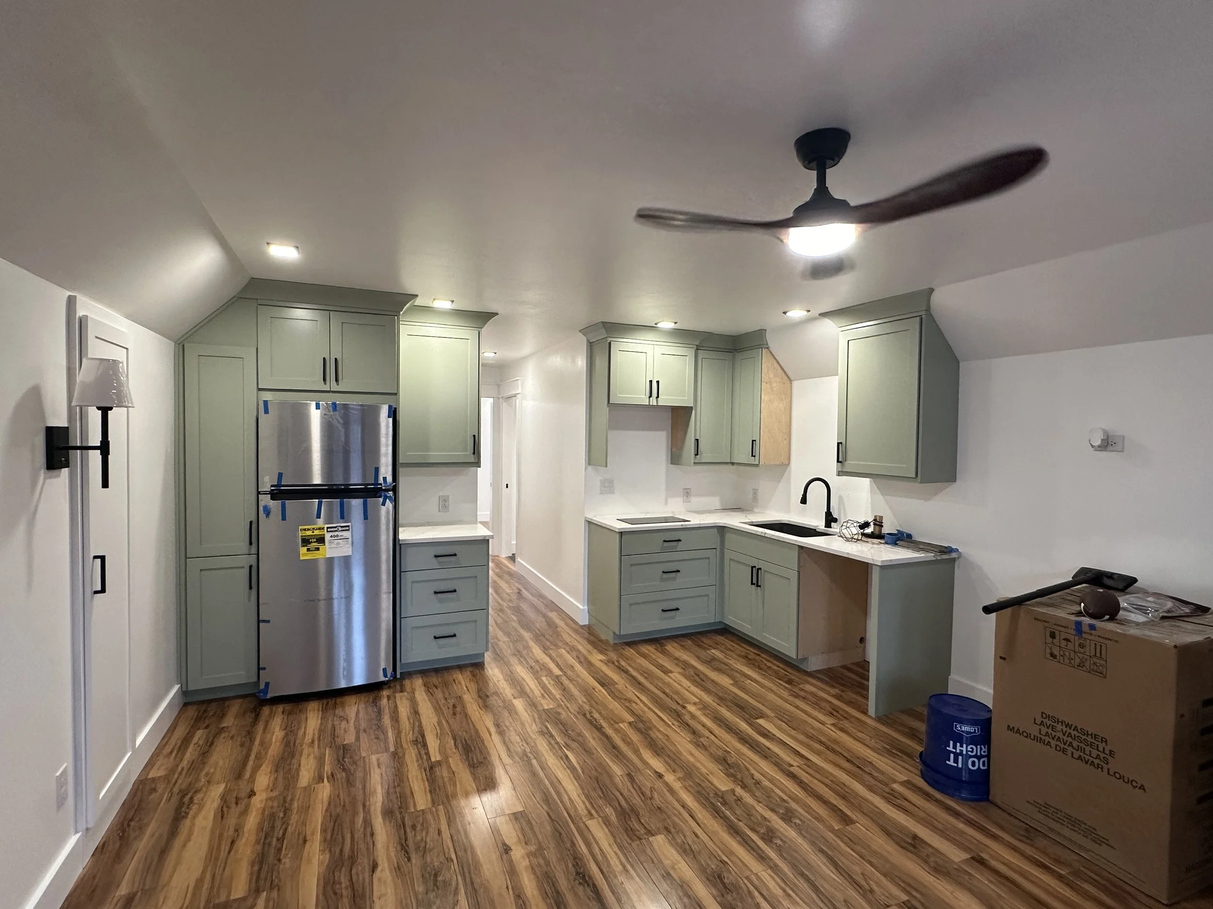 A kitchen under construction with green cabinets, a stainless steel refrigerator, a black faucet, and a ceiling fan with lights. There are boxes and a blue container on the wooden floor.