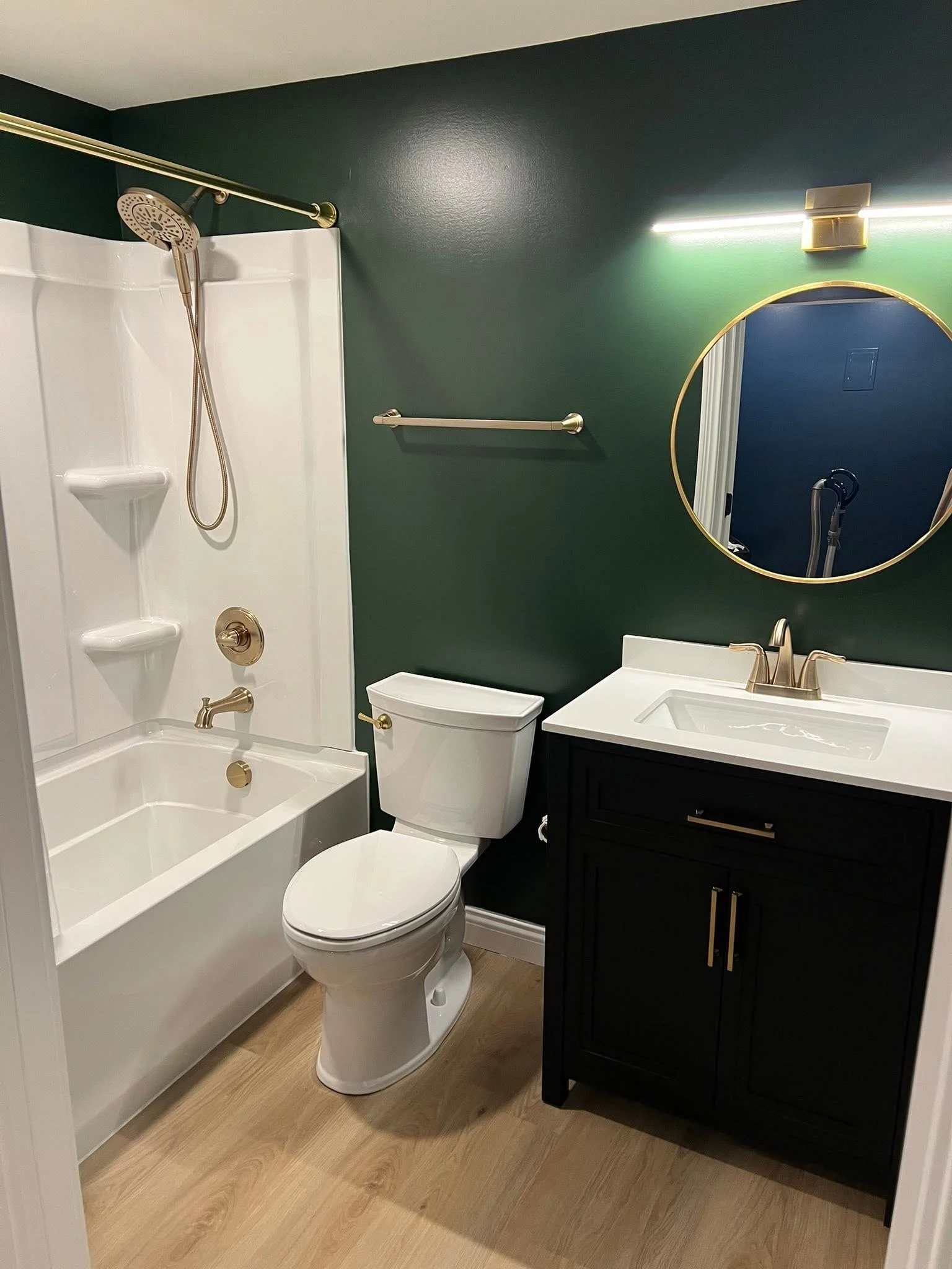 A modern bathroom featuring a bathtub with a shower and a gold showerhead, a toilet, a black vanity with a white sink, a round mirror with a gold frame, and dark green walls with light wood flooring.