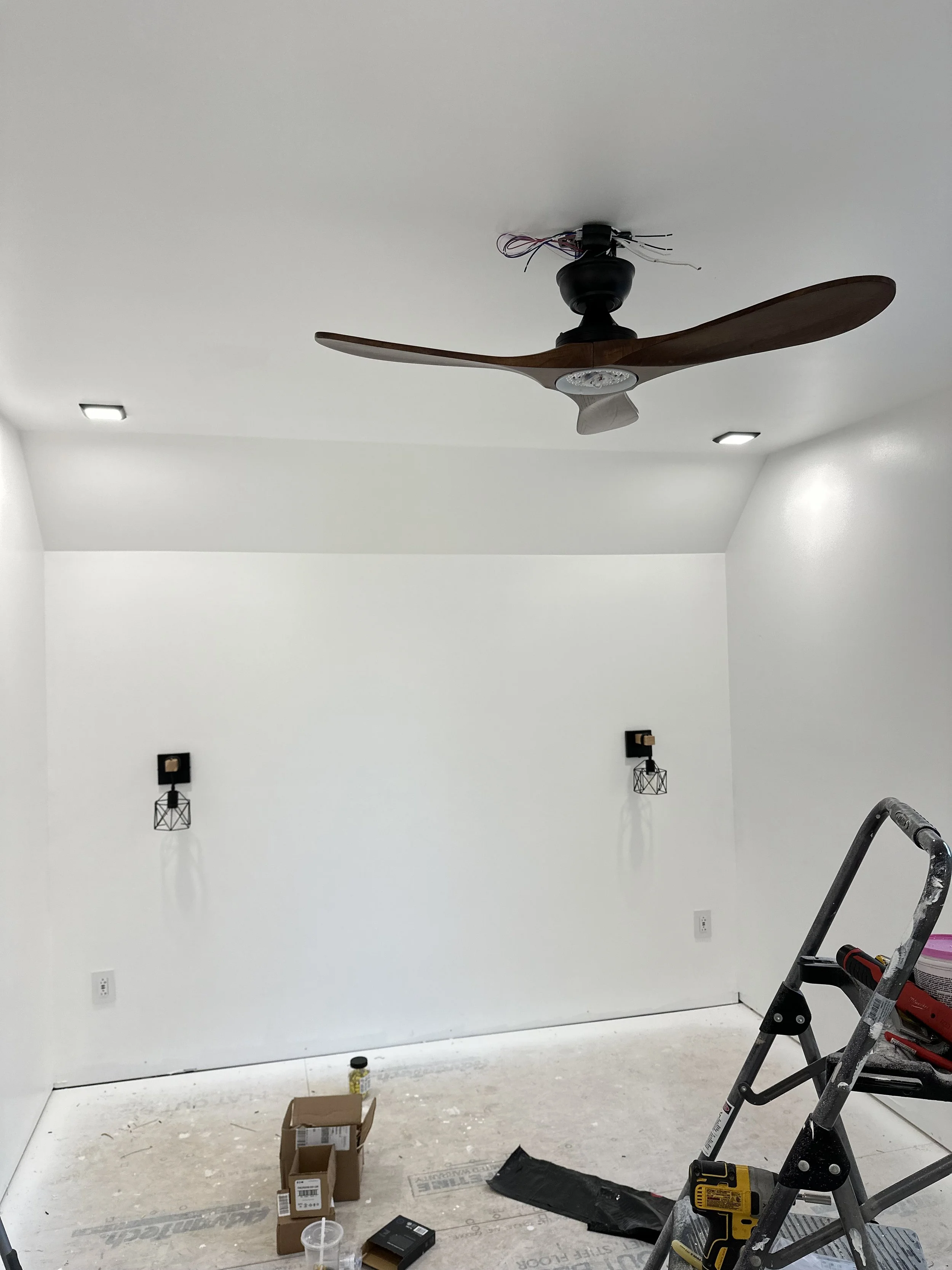 A room with white walls and ceiling, under construction or renovation. Ceiling has no light fixture, wiring exposed. On the wall are two black wall-mounted light fixtures with cage covers. The floor is unfinished with tools, boxes, and a ladder prese