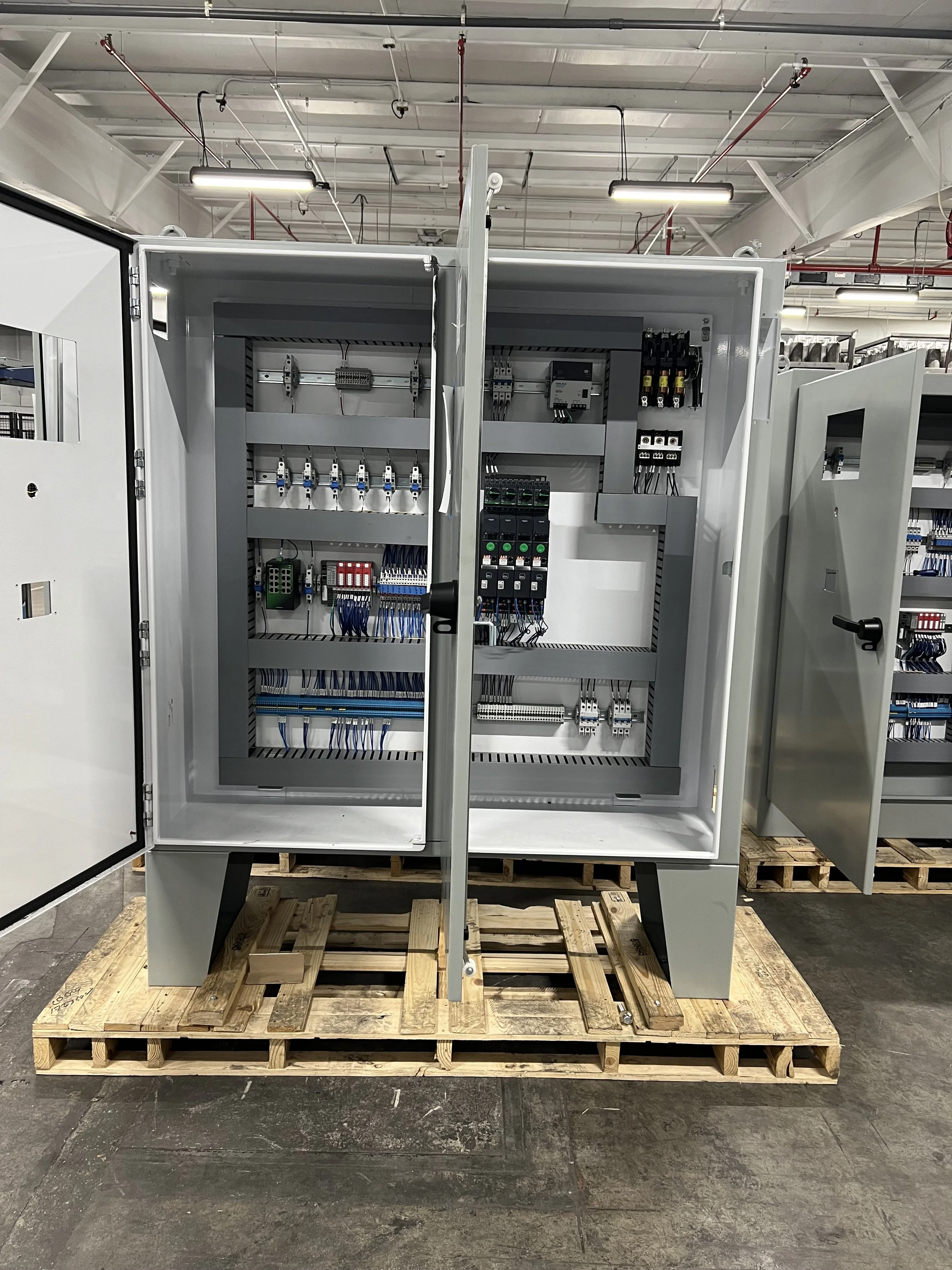 An industrial electrical control cabinet with various wires, circuit breakers, and components inside, placed on a wooden pallet in a warehouse or factory setting.